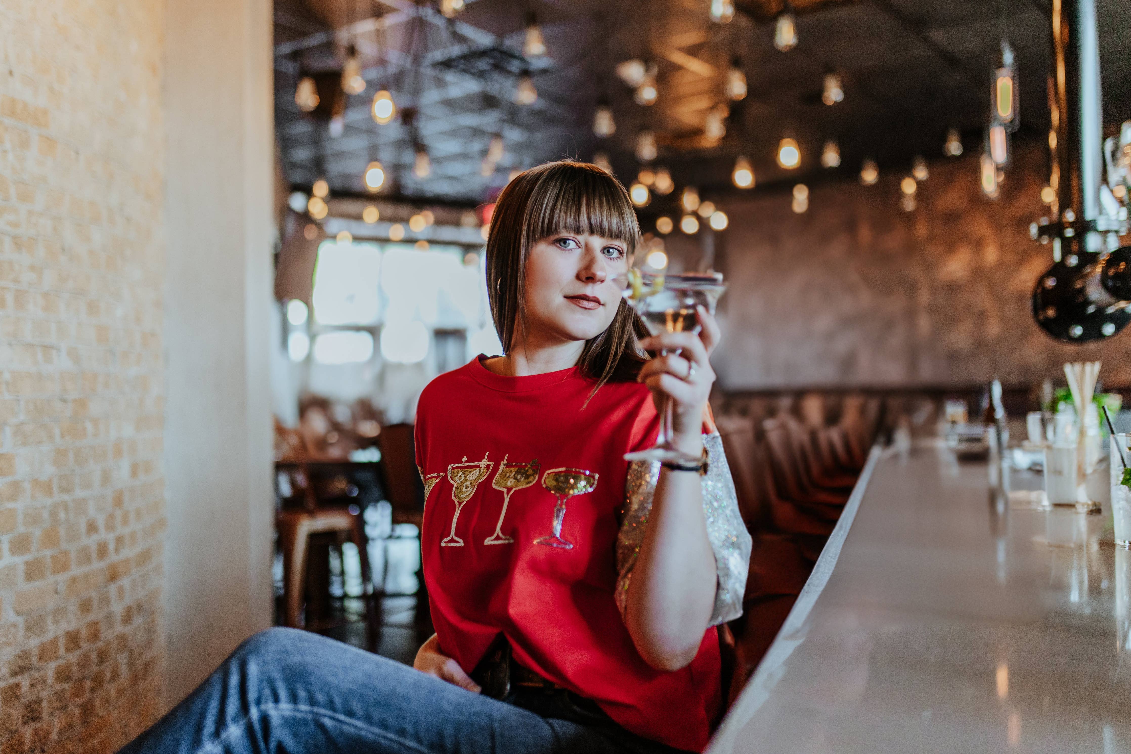 Person in a red shirt with drink designs sitting at a bar counter.