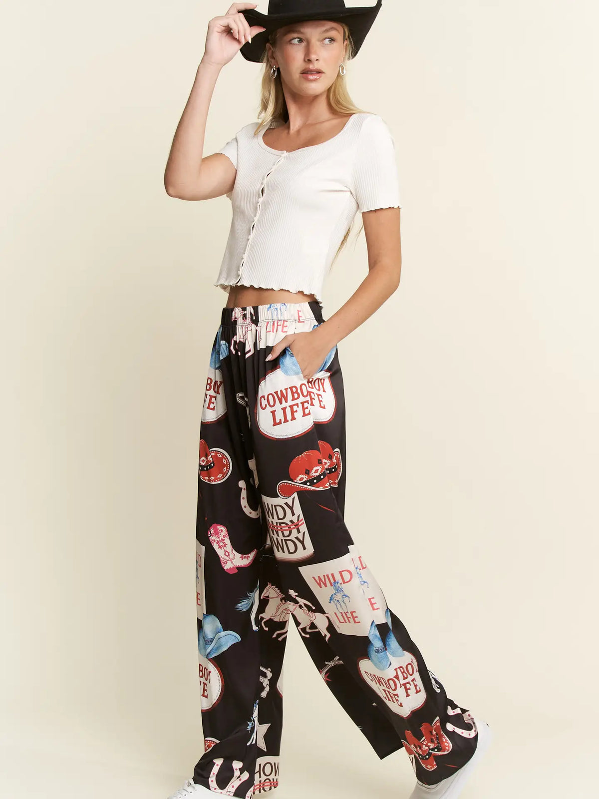 A woman modeling a white cropped short-sleeve top and black wide-leg pants with a colorful western-themed graphic print, including a cowboy hat, against a plain beige studio background.