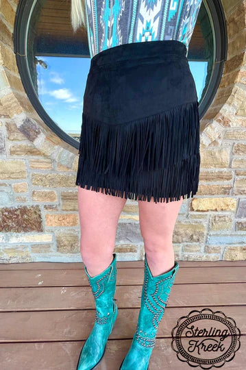 A person wearing a black fringe mini skirt and turquoise western cowboy boots, standing on a wooden deck in front of a stone wall and a round mirror. A 'Sterling Kreek' logo watermark is visible in the bottom right corner.