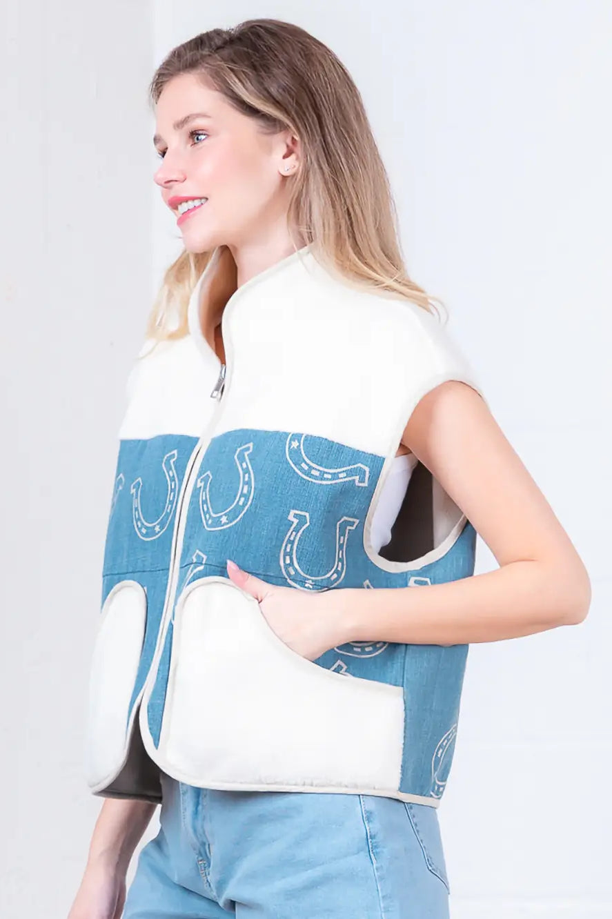 Model wearing indigo denim vest with white horseshoe print and faux-fur collar, zipped front, hands in pockets.”