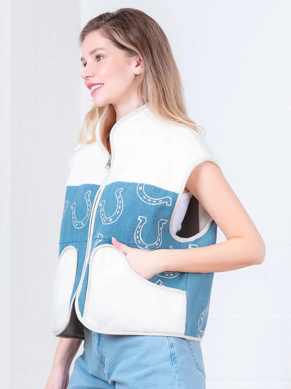 Model wearing indigo denim vest with white horseshoe print and faux-fur collar, zipped front, hands in pockets.”