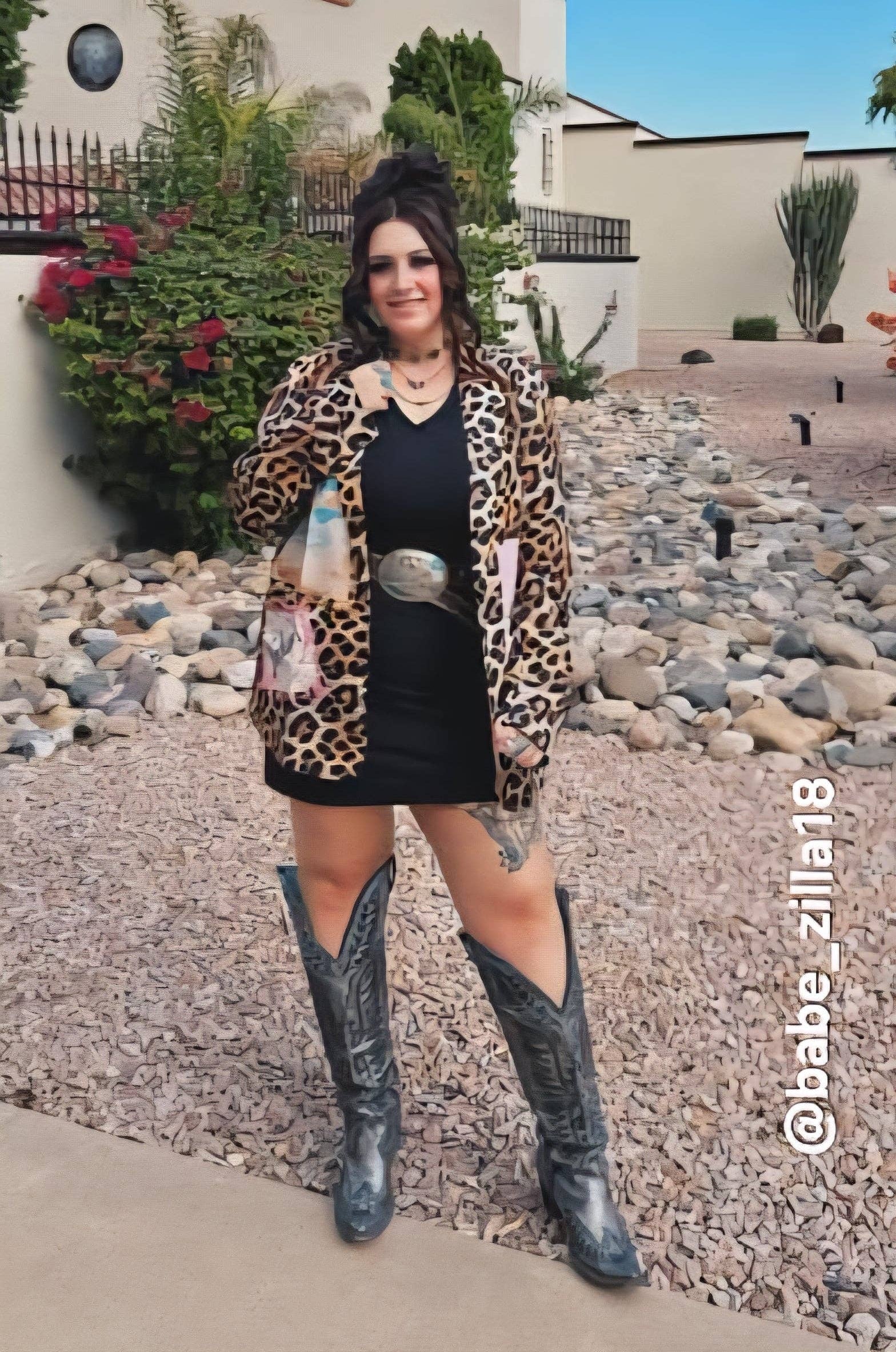 Woman in leopard print cardigan and black dress with cowboy boots standing in front of a building.
