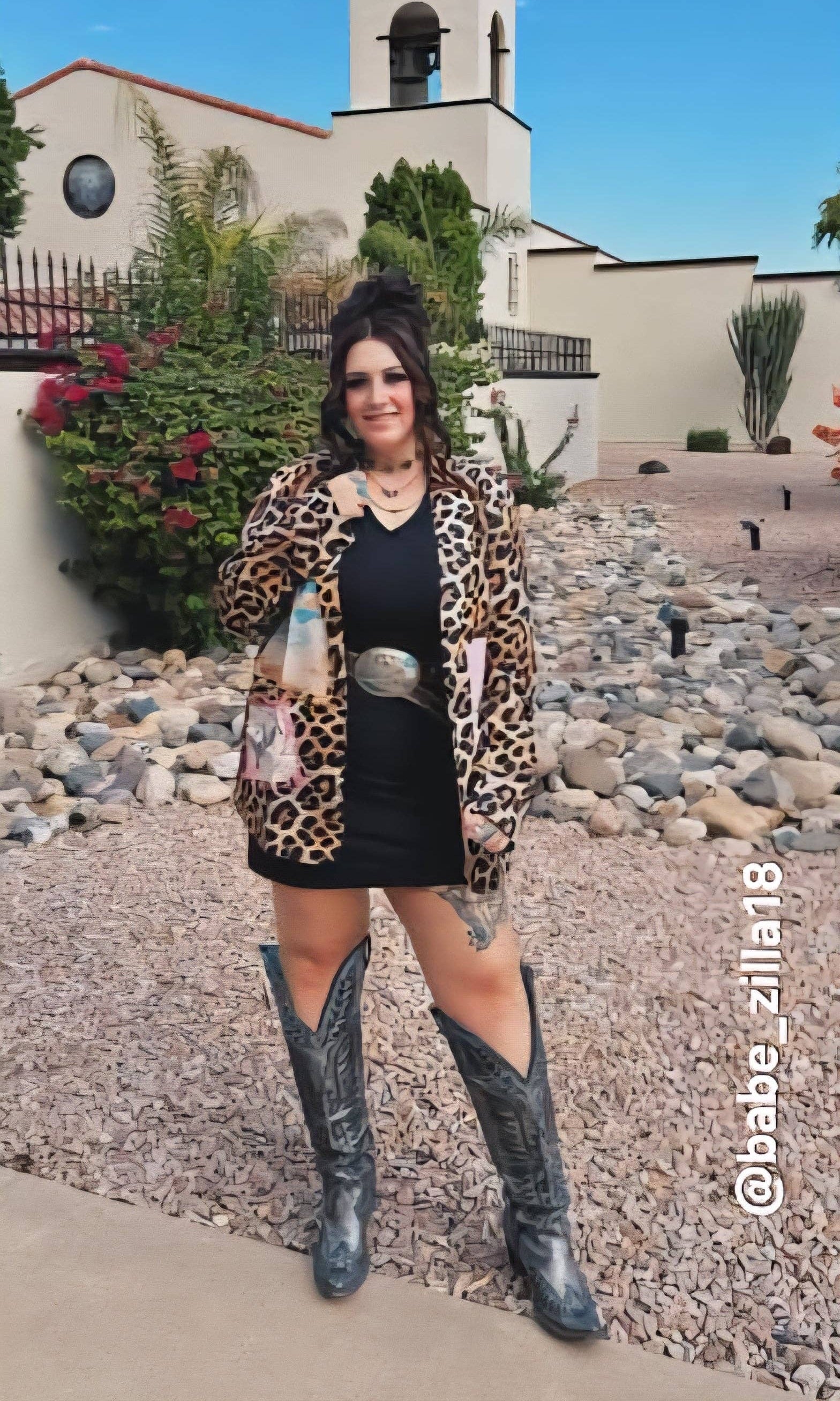 Woman in leopard print cardigan and black dress with cowboy boots standing in front of a building.