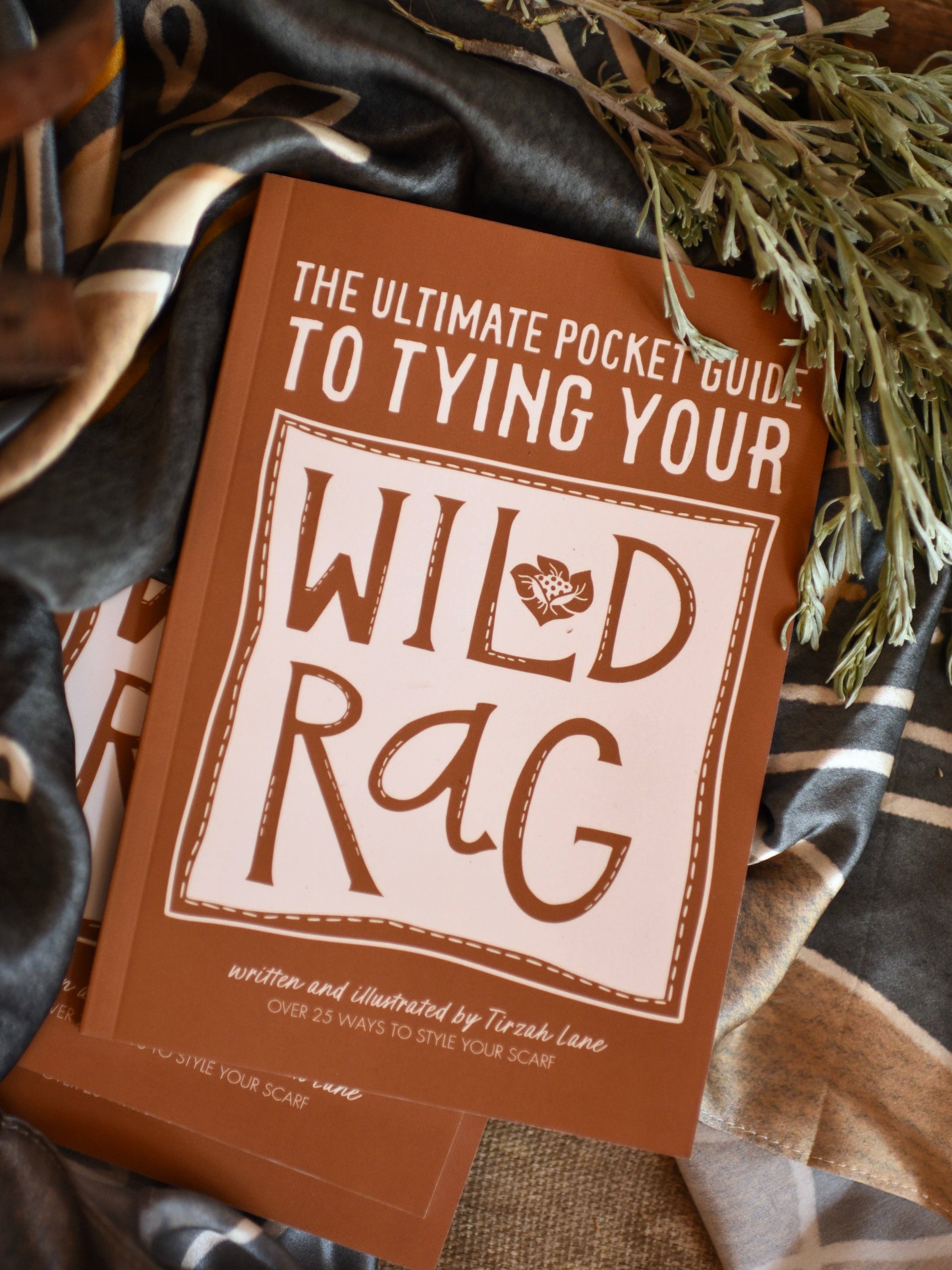 Book titled 'The Ultimate Pocket Guide to Tying Your Wild Rag' on a textured surface with fabric and plants.