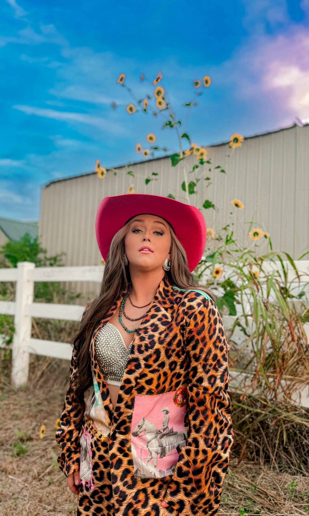 Woman in a leopard print outfit and pink hat standing in a field with sunflowers and a barn in the background.