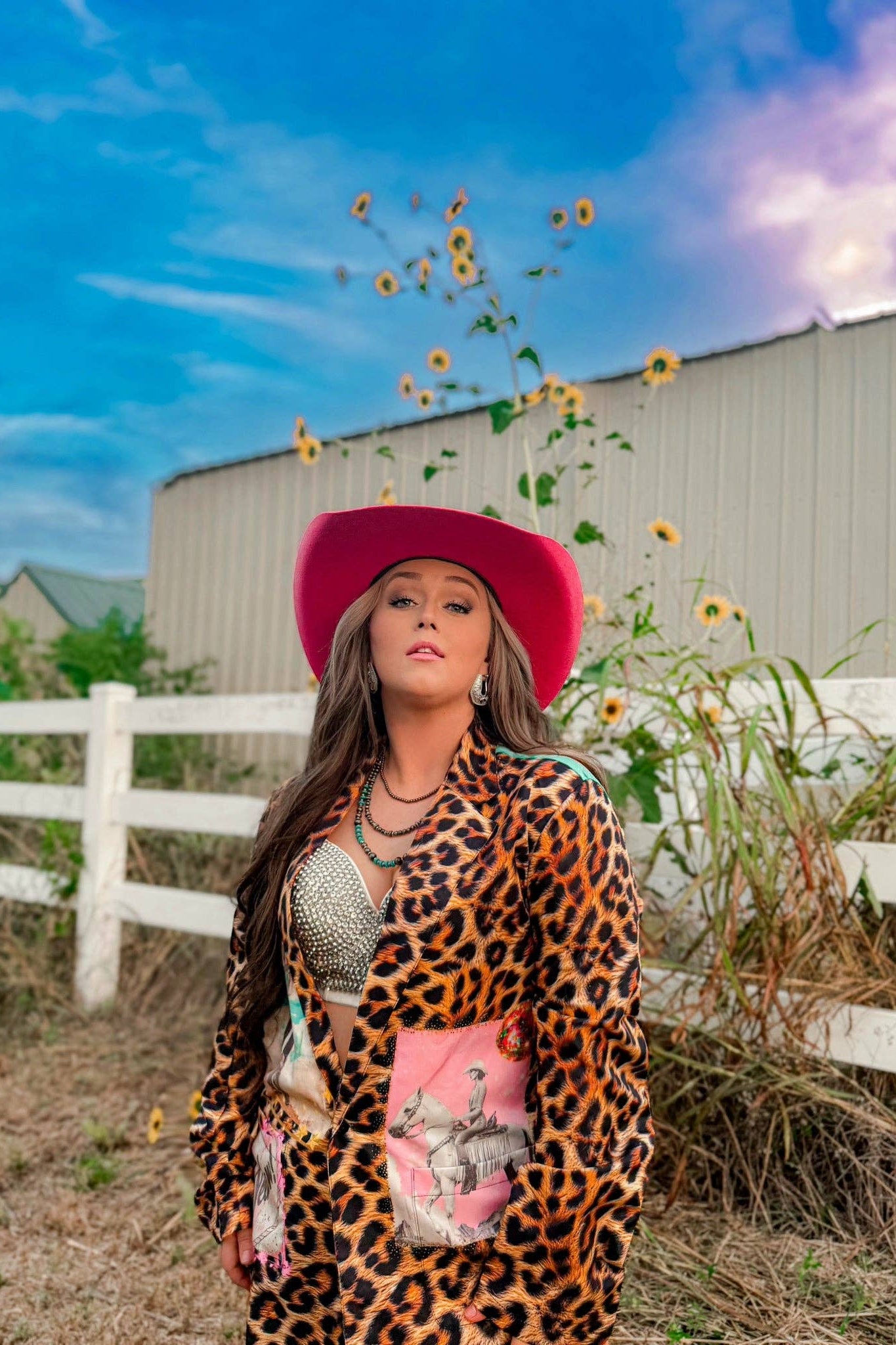 Woman in a leopard print outfit and pink hat standing in a field with sunflowers and a barn in the background.