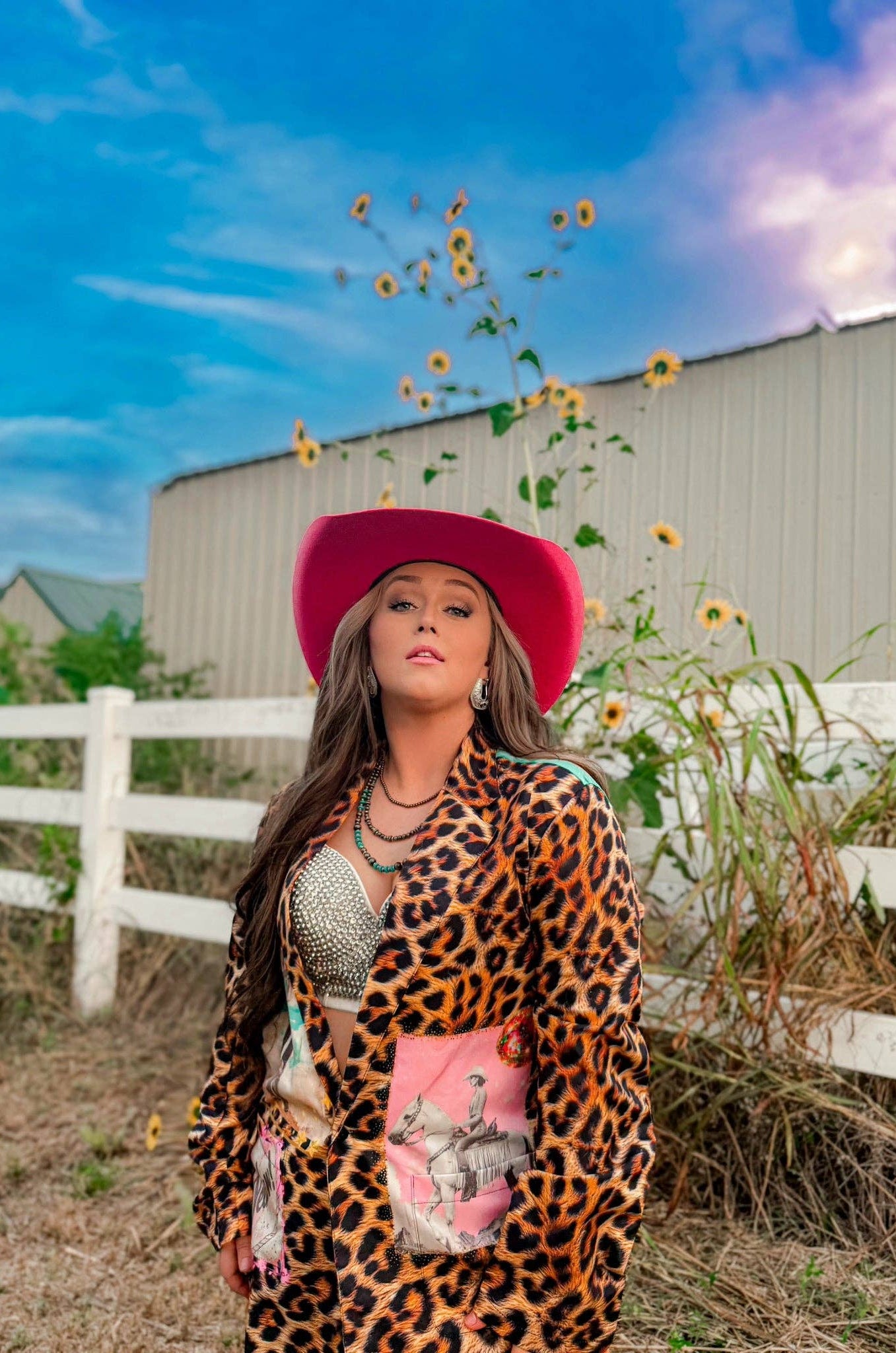 Woman in a leopard print outfit and pink hat standing in a field with sunflowers and a barn in the background.