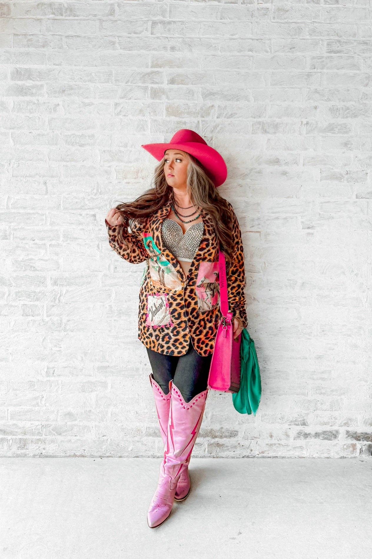 Person wearing a leopard print cardigan, pink cowboy hat, pink boots, and colorful handbag against a white brick wall.