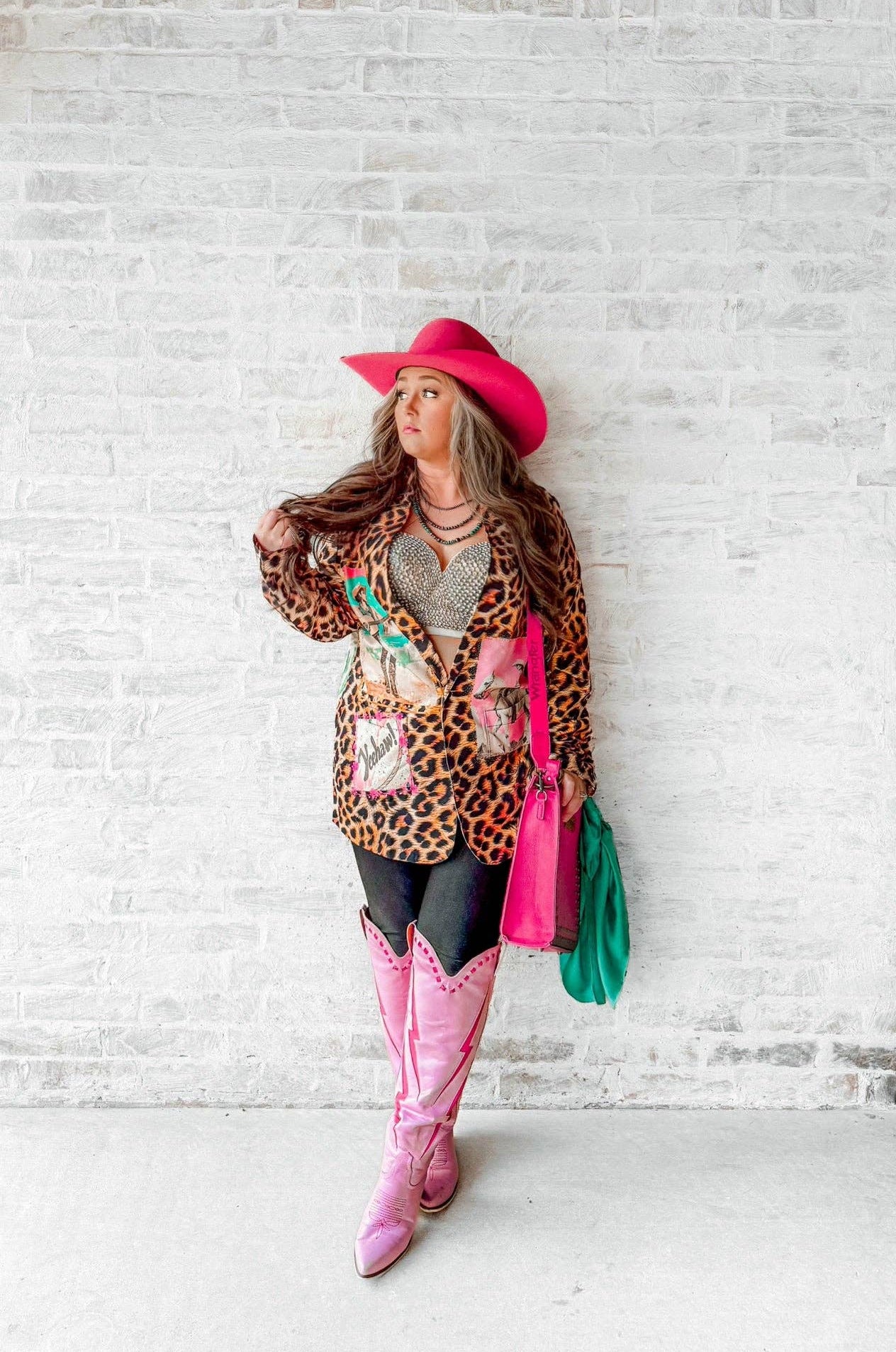 Person wearing a leopard print cardigan, pink cowboy hat, pink boots, and colorful handbag against a white brick wall.