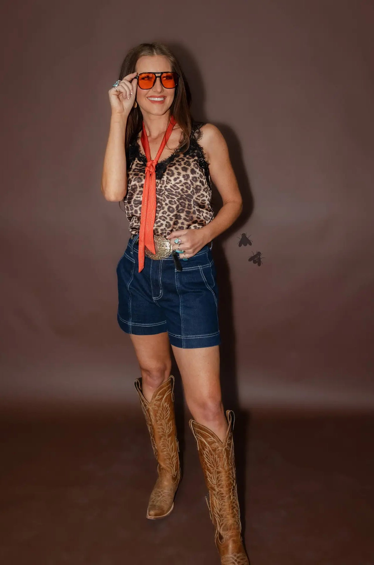 Woman wearing a leopard print top, denim shorts, and cowboy boots against a brown background