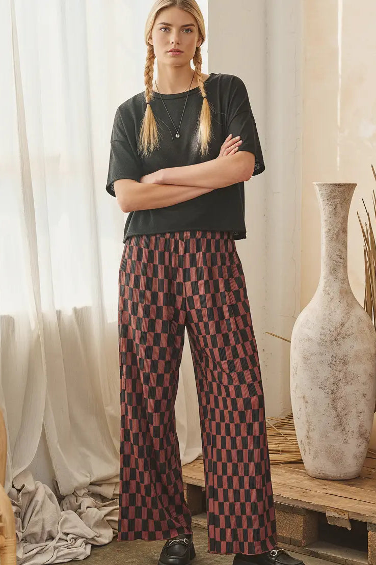A woman stands indoors with arms crossed, wearing a black short-sleeve top and wide-leg trousers with a red and black checkered pattern. The setting includes sheer curtains, a rustic wooden bench, large ceramic vases, and dried plant stems.