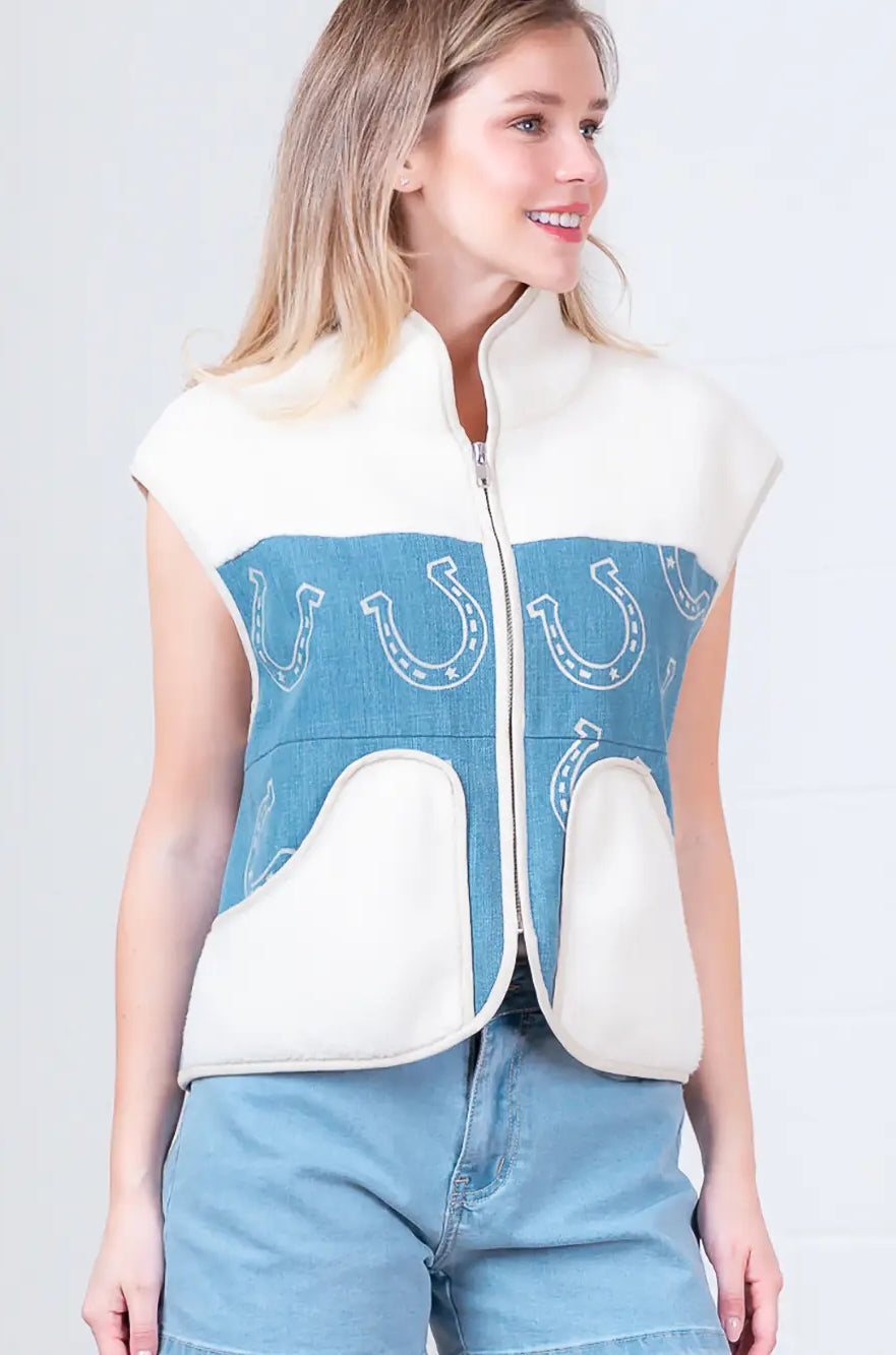 Model wearing indigo denim vest with white horseshoe print and faux-fur collar, zipped front, hands in pockets.”