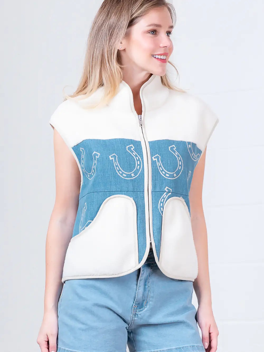 Model wearing indigo denim vest with white horseshoe print and faux-fur collar, zipped front, hands in pockets.”