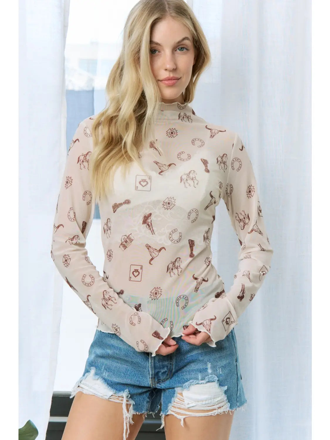 A woman modeling a beige long-sleeve mock-neck top with a brown bohemian-style pattern, paired with distressed denim shorts, set against a soft white curtain background.