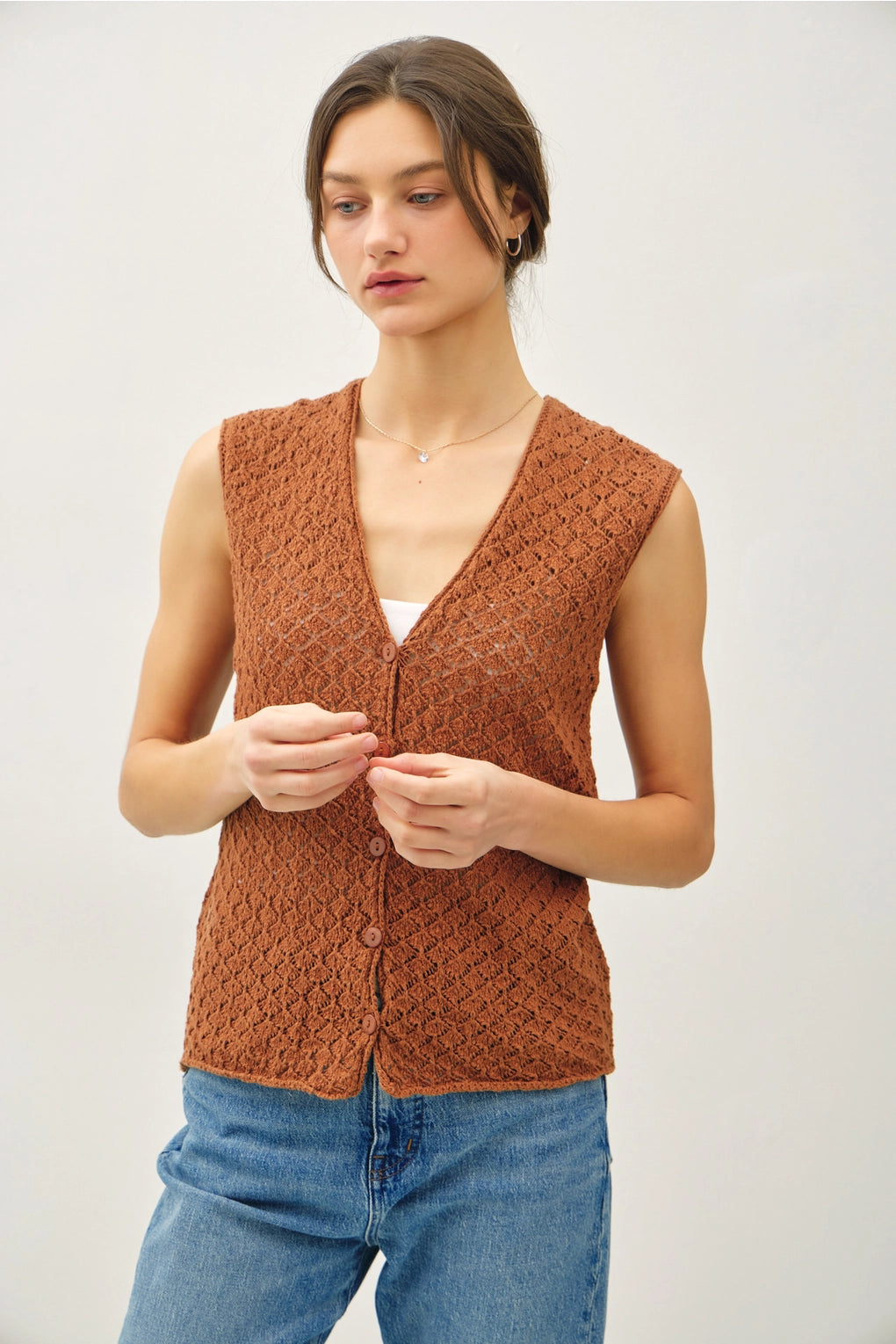 Woman wearing a brown knitted vest over a white shirt and blue jeans on a light background