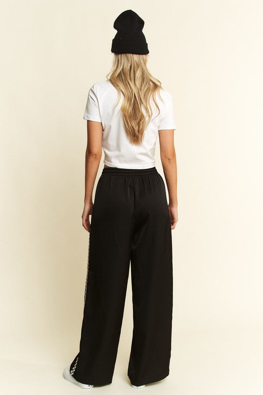 A woman shown from the back wearing a black beanie, white t-shirt, and black wide-leg pants with white side stripes, standing against a plain cream background.