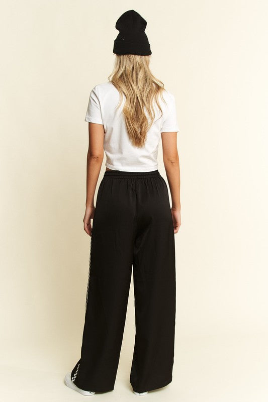 A woman shown from the back wearing a black beanie, white t-shirt, and black wide-leg pants with white side stripes, standing against a plain cream background.