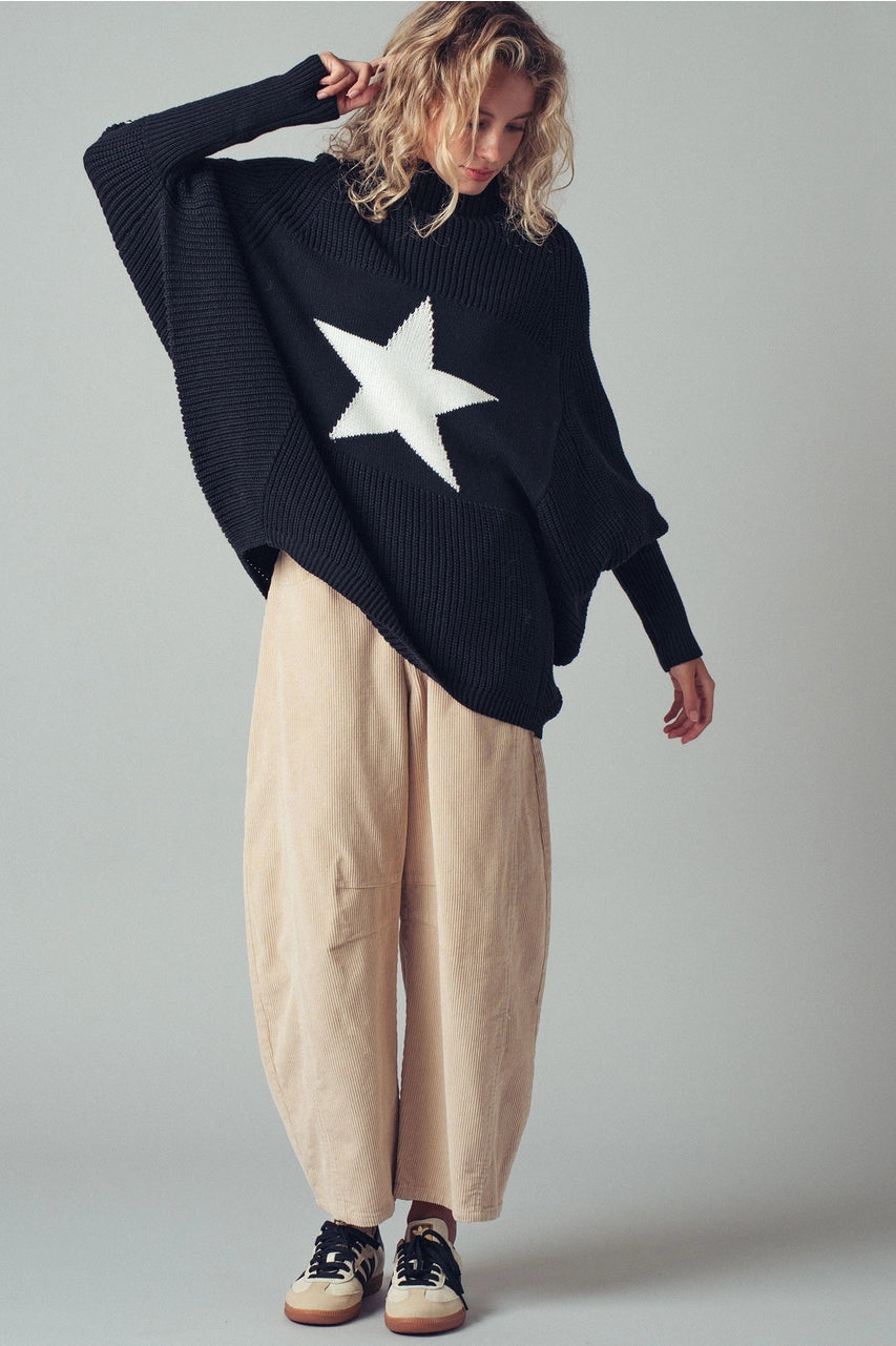Person wearing a black sweater with a white star and beige pants on a plain background