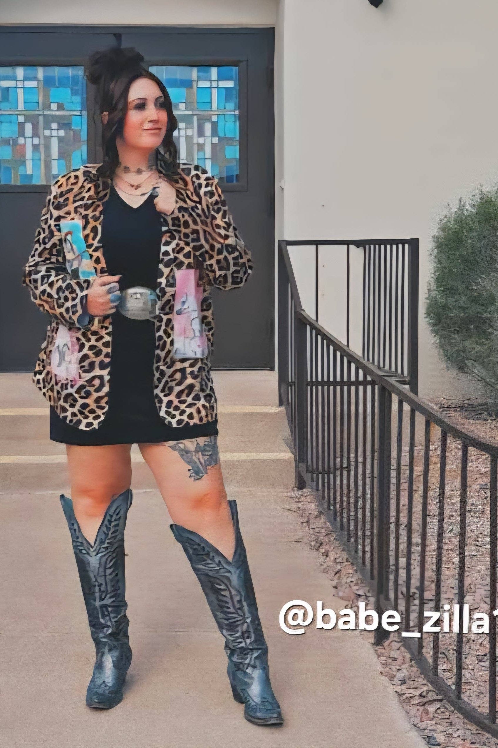 Person wearing a leopard print jacket, black dress, and blue cowboy boots standing outside.