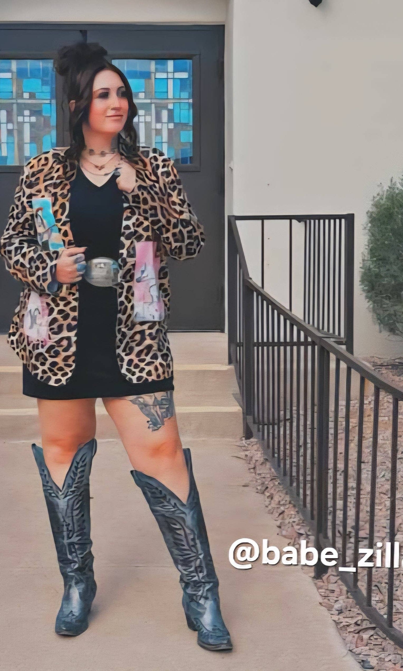 Person wearing a leopard print jacket, black dress, and blue cowboy boots standing outside.