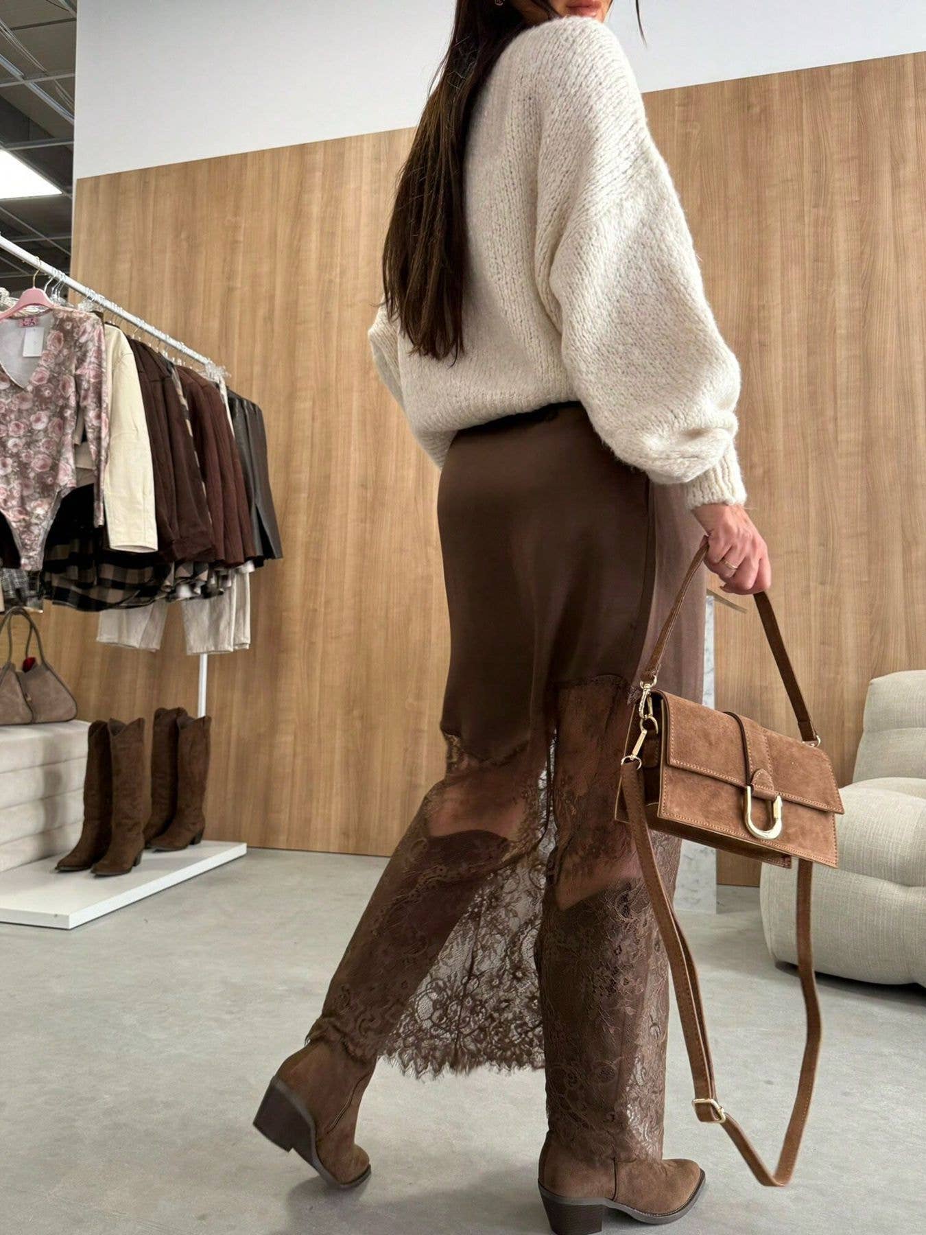 Person wearing a white sweater and brown pants holding a brown handbag in a store setting.