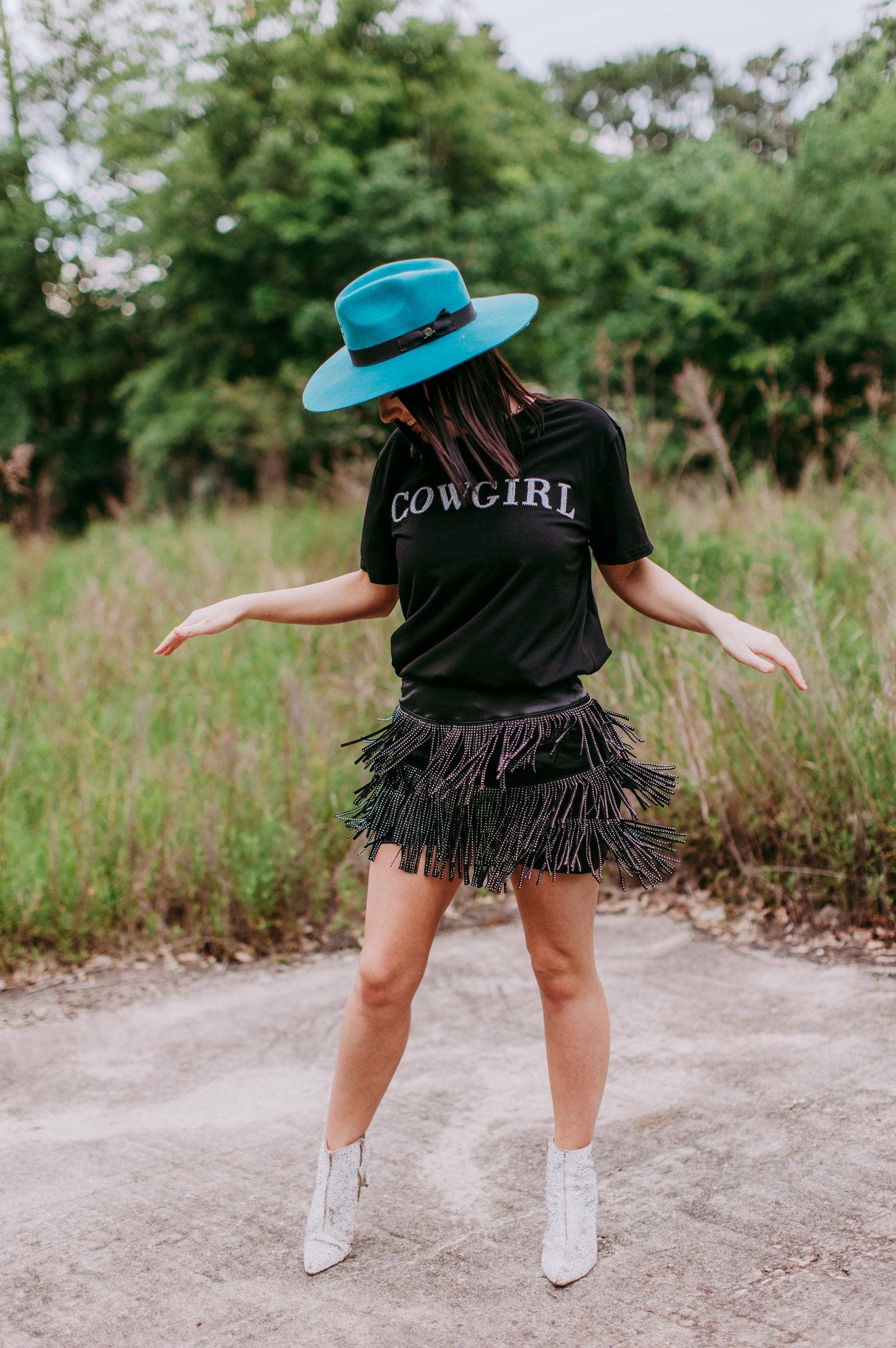 Person wearing a black 'COWGIRL' shirt, fringed skirt, and blue hat outdoors.