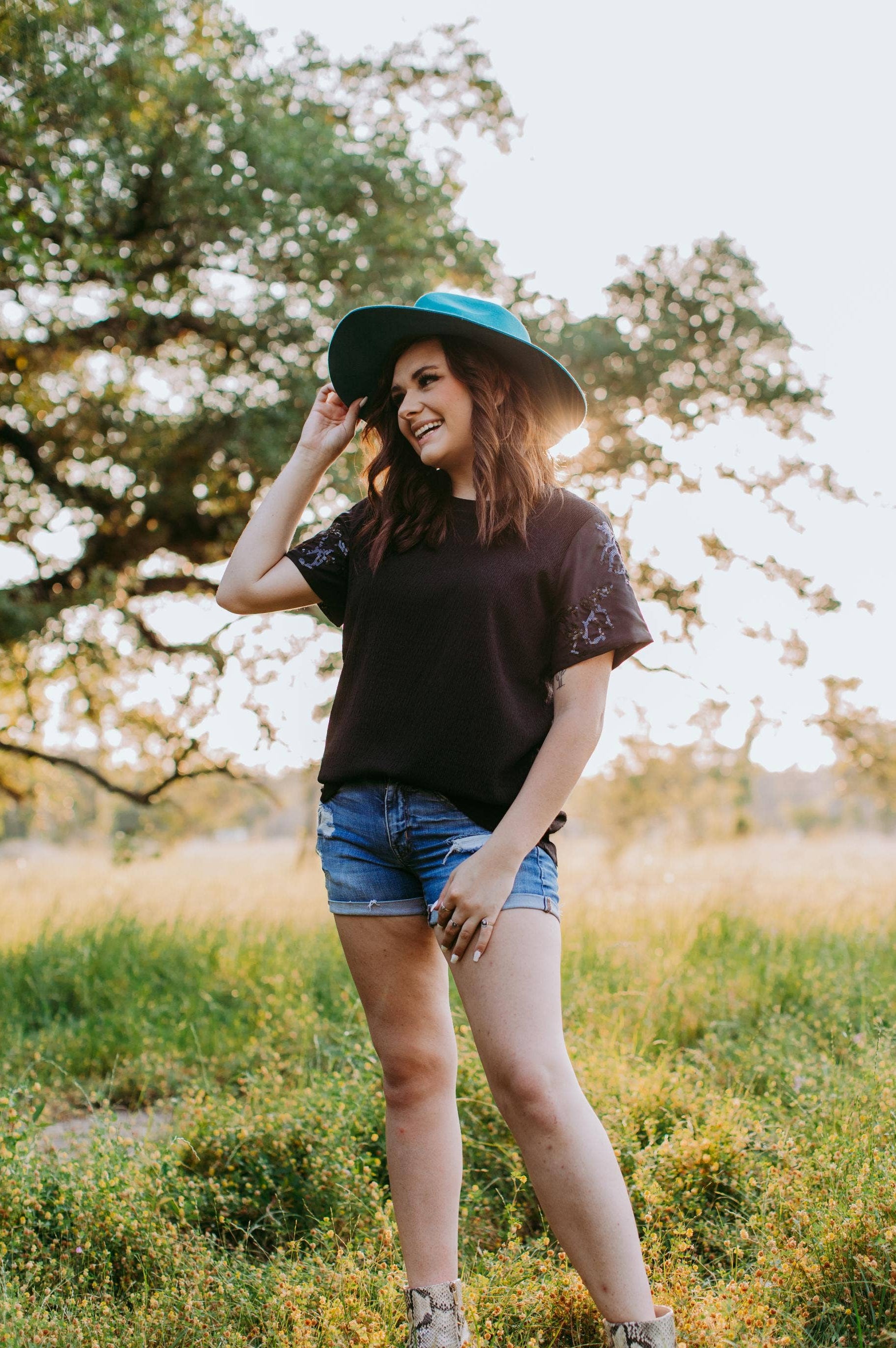 Woman wearing a black t-shirt, denim shorts, and a blue hat standing in a field with trees in the background.