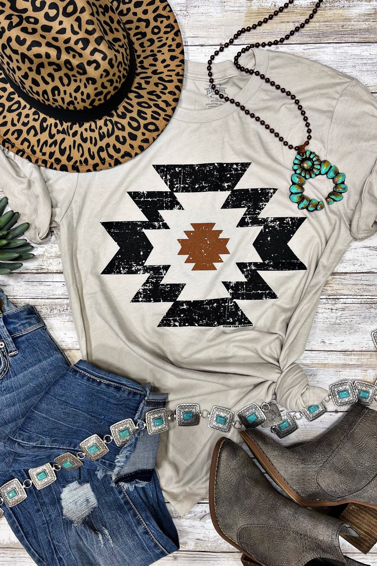 White t-shirt with black geometric pattern, leopard print hat, blue jeans, and brown boots on a wooden surface.