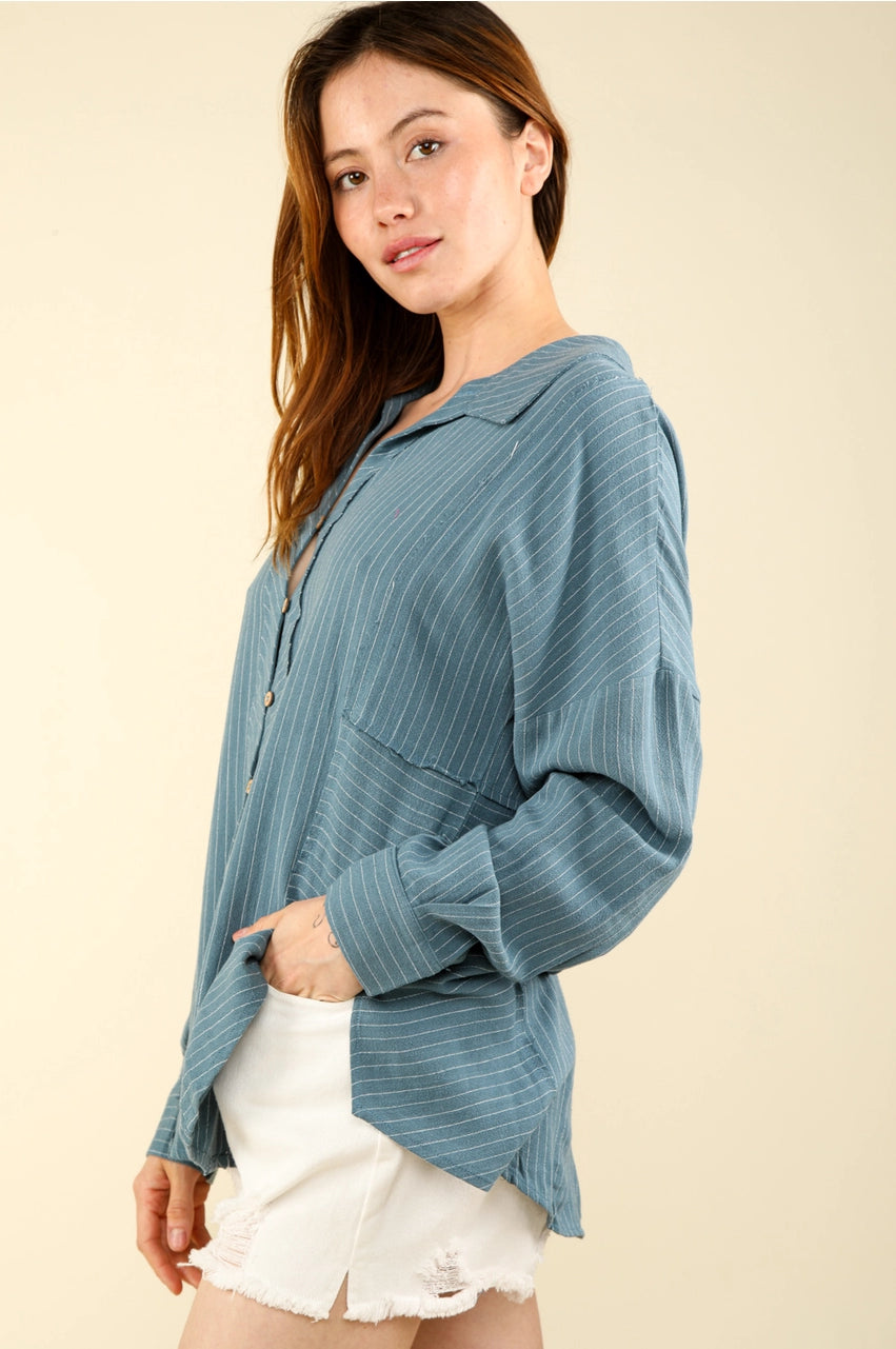 Side view of dusty-blue striped button-up shirt with drop shoulder and long sleeves; model in white denim shorts.”