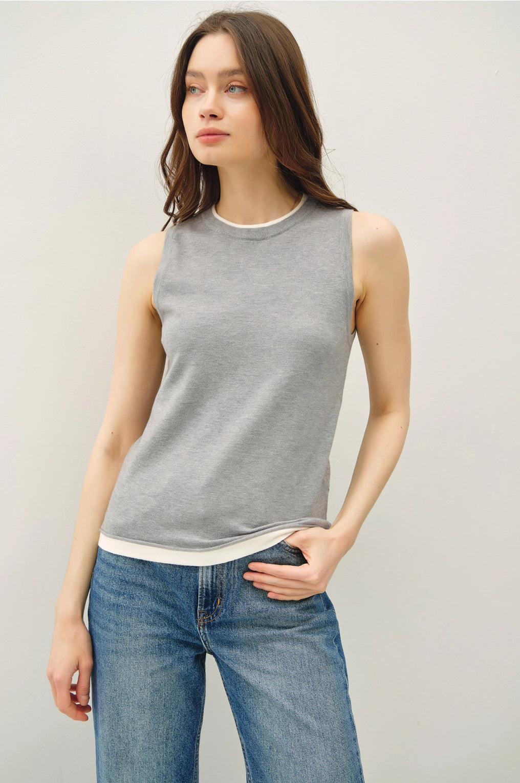 Woman wearing a light grey sleeveless top with lighter contrasting band at the bottom of the tops and blue jeans on a plain background