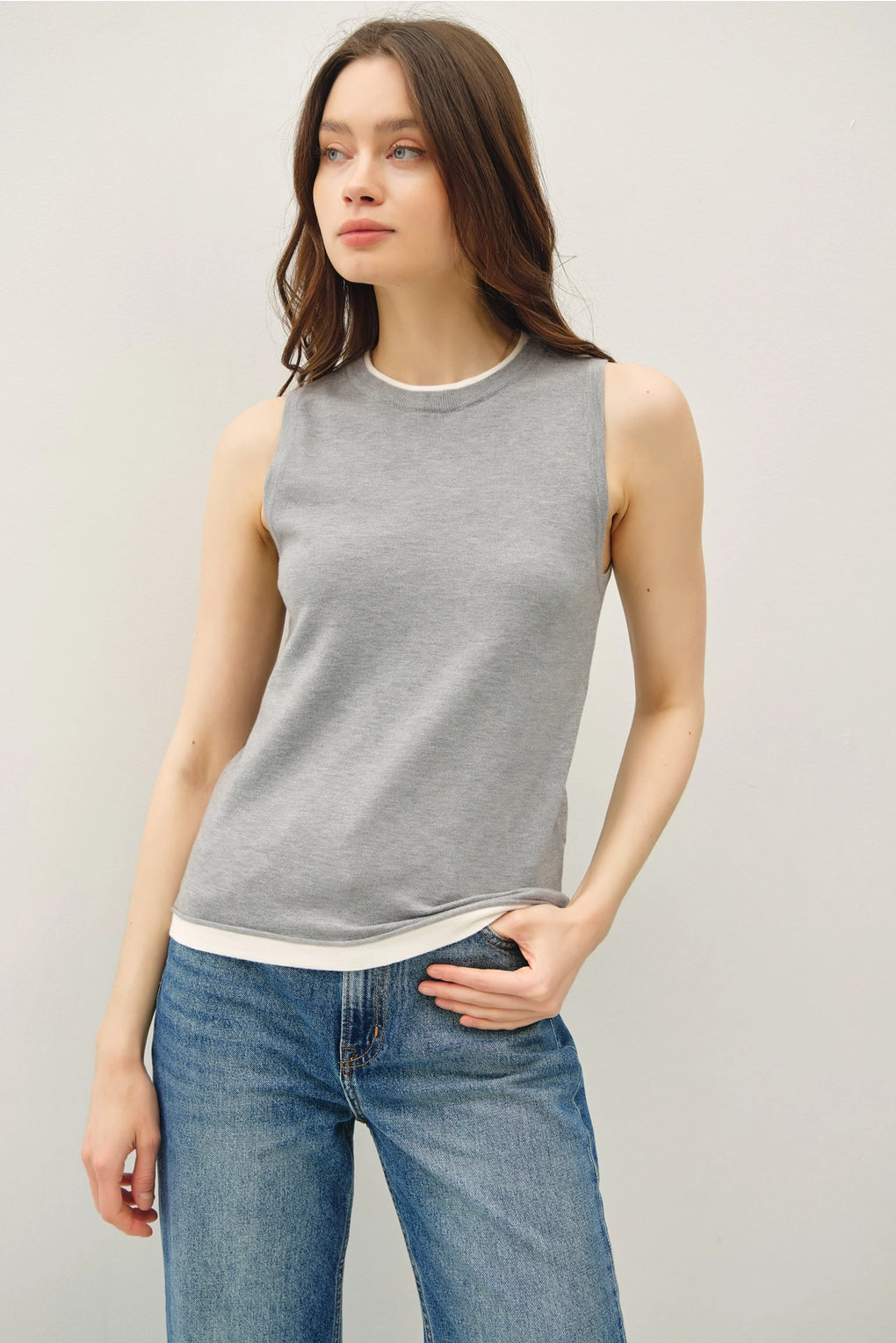 Woman wearing a light grey sleeveless top with lighter contrasting band at the bottom of the tops and blue jeans on a plain background