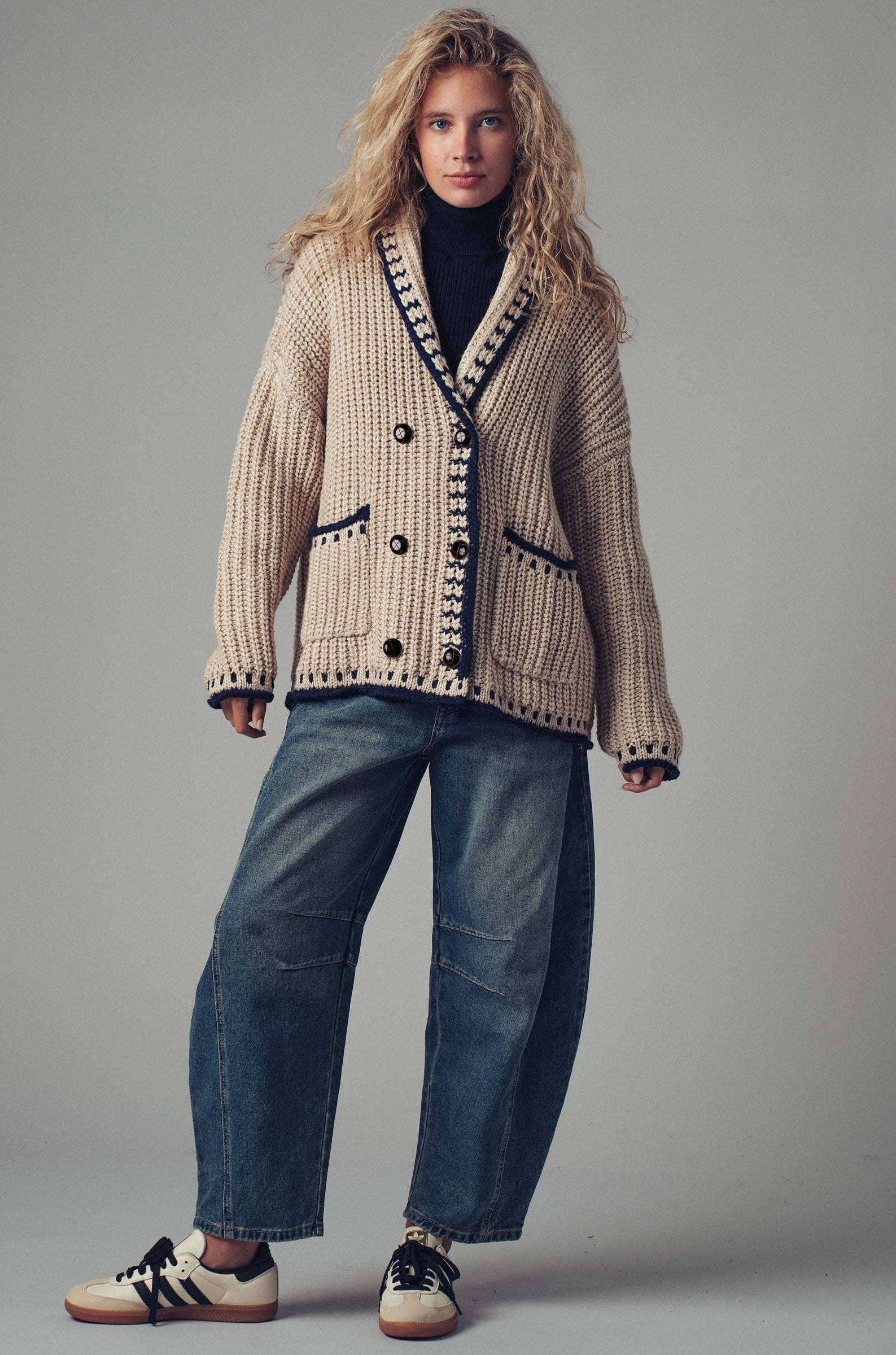 Woman wearing a patterned cardigan, black turtleneck, blue jeans, and sneakers on a gray background