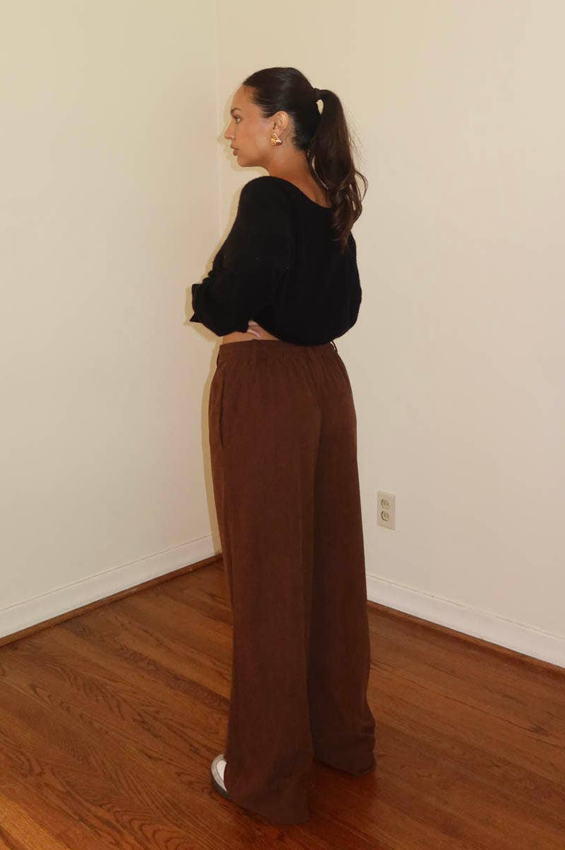 Person wearing black top and brown wide-leg pants in a room with wooden floor and white walls.