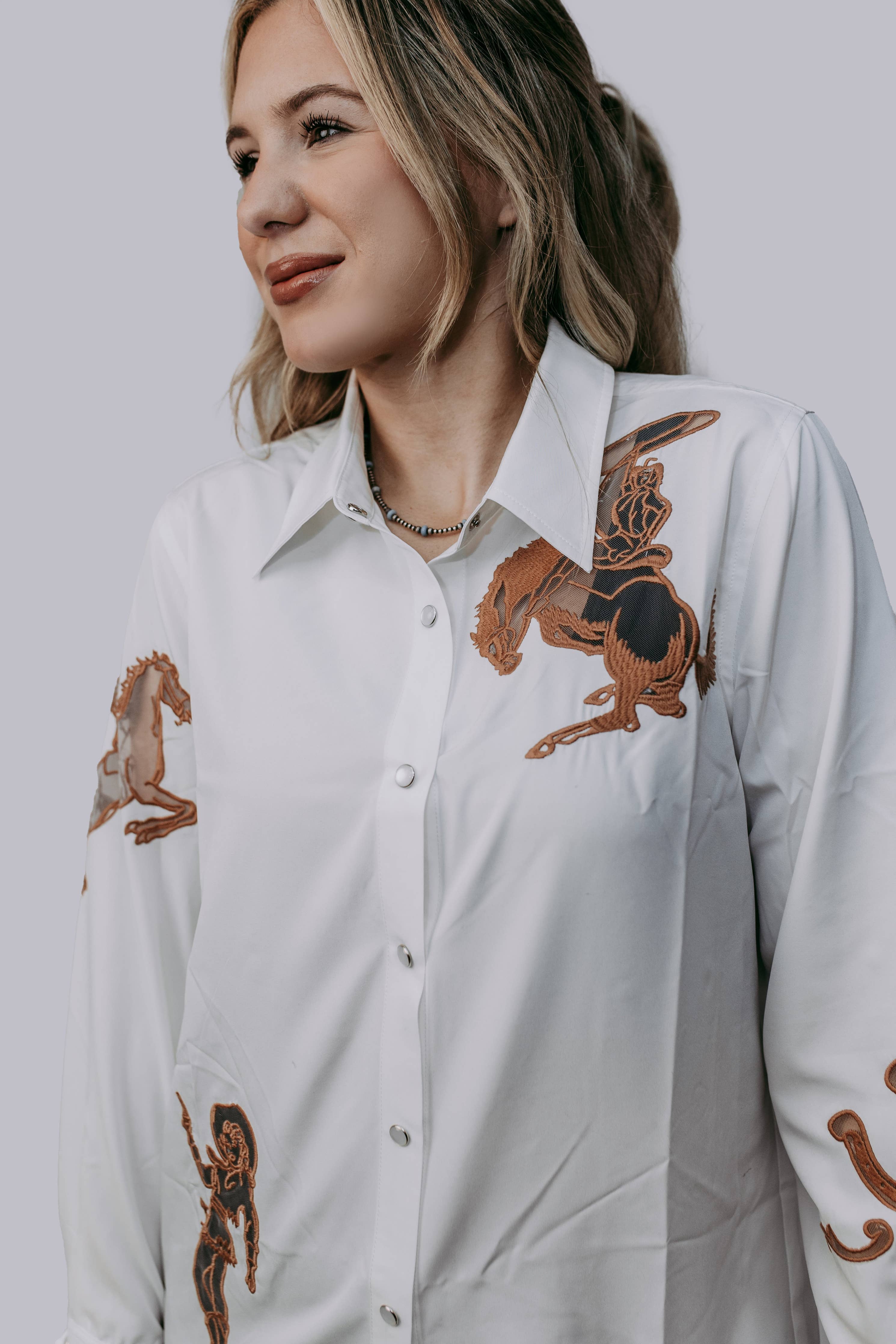 A woman smiling and posing in a white button-down blouse with cut out embroidery western horseshoes and cowboys with pearl snap buttons, paired with dark pants.