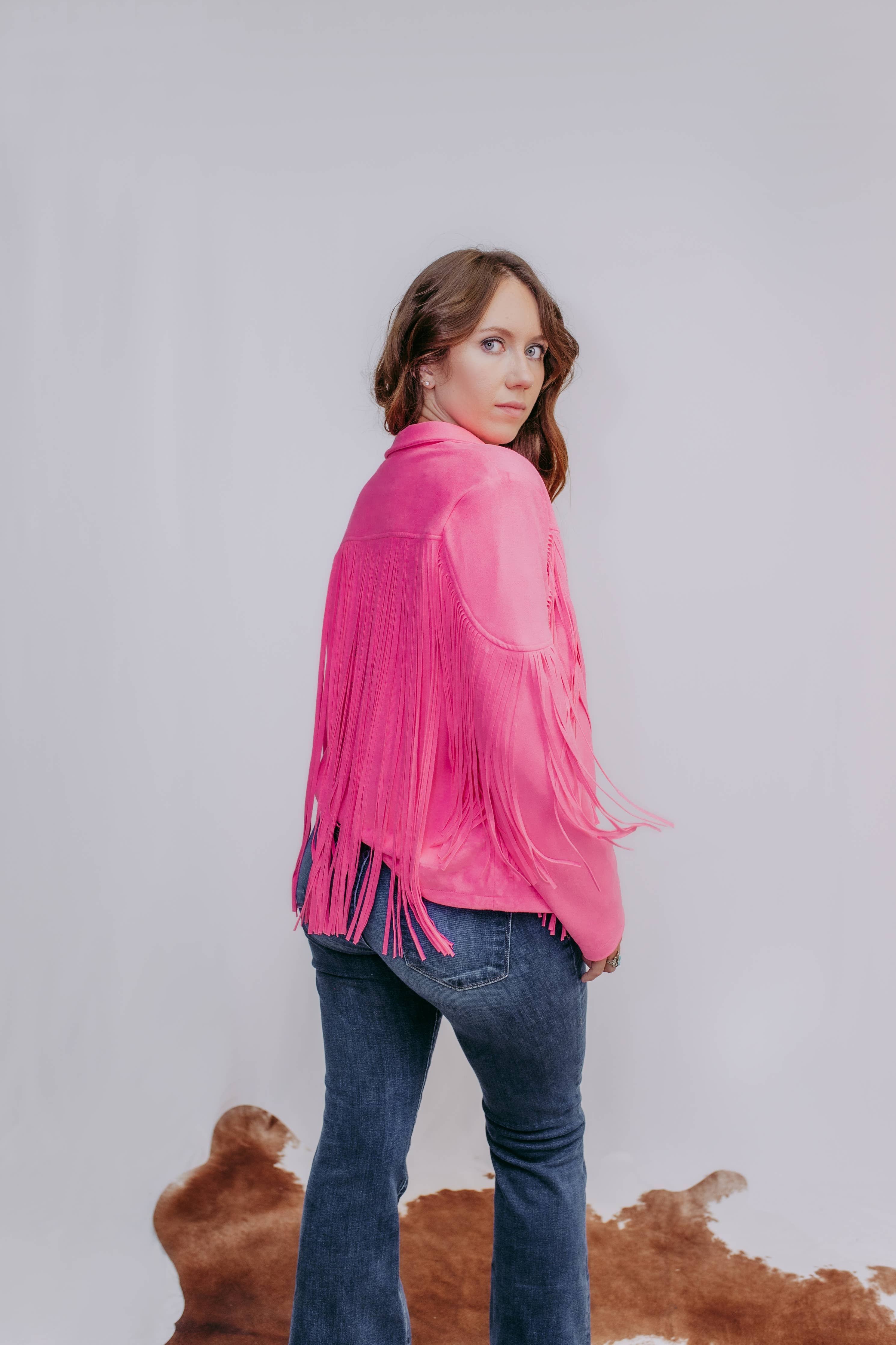 Woman wearing a pink fringed jacket and blue jeans against a plain background