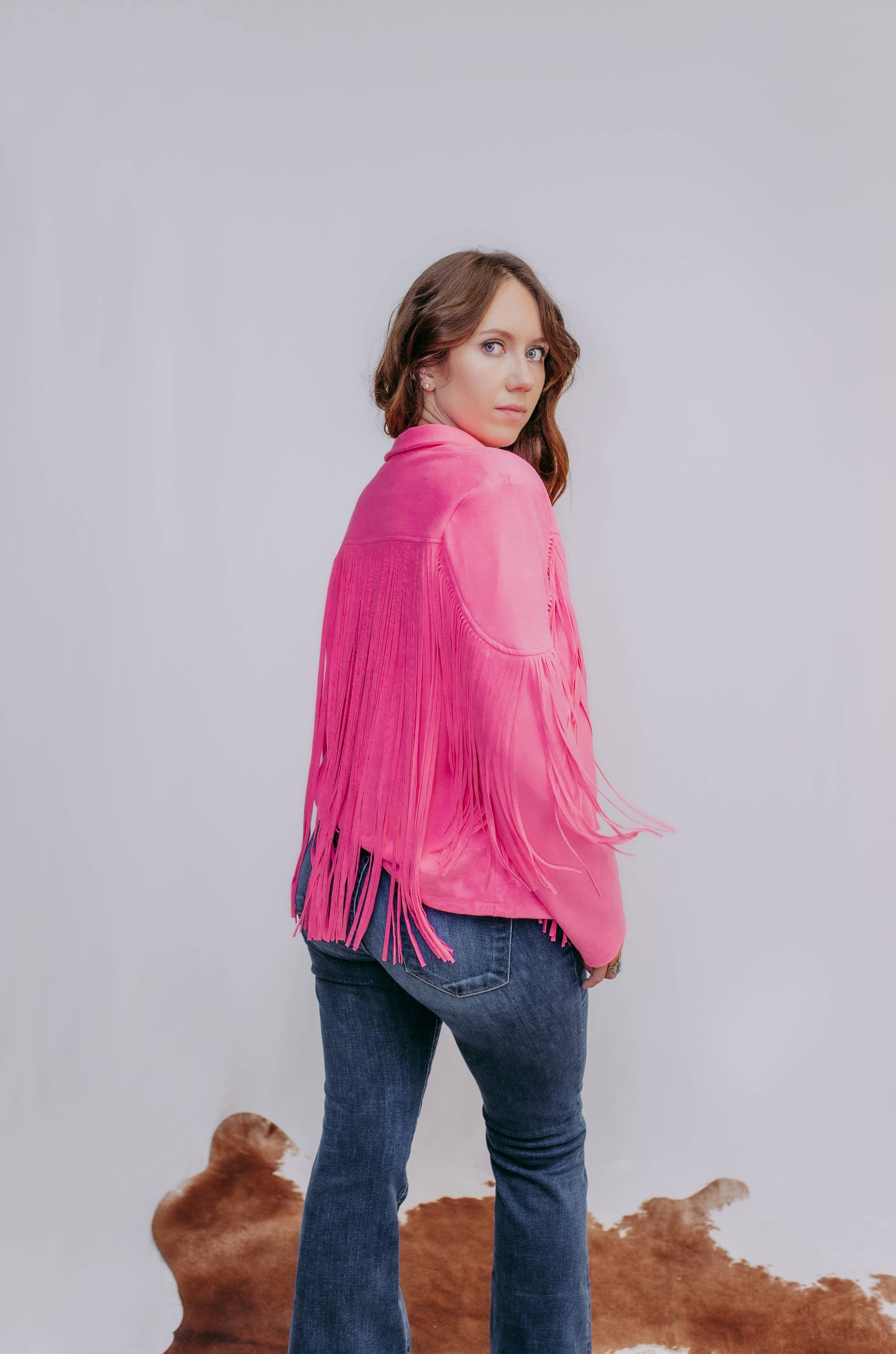 Woman wearing a pink fringed jacket and blue jeans against a plain background