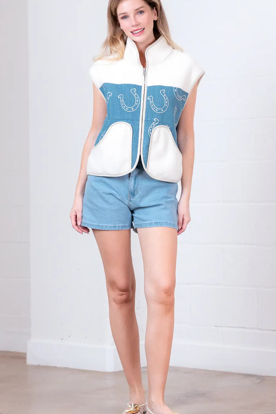 Model wearing indigo denim vest with white horseshoe print and faux-fur collar, zipped front, hands in pockets.”