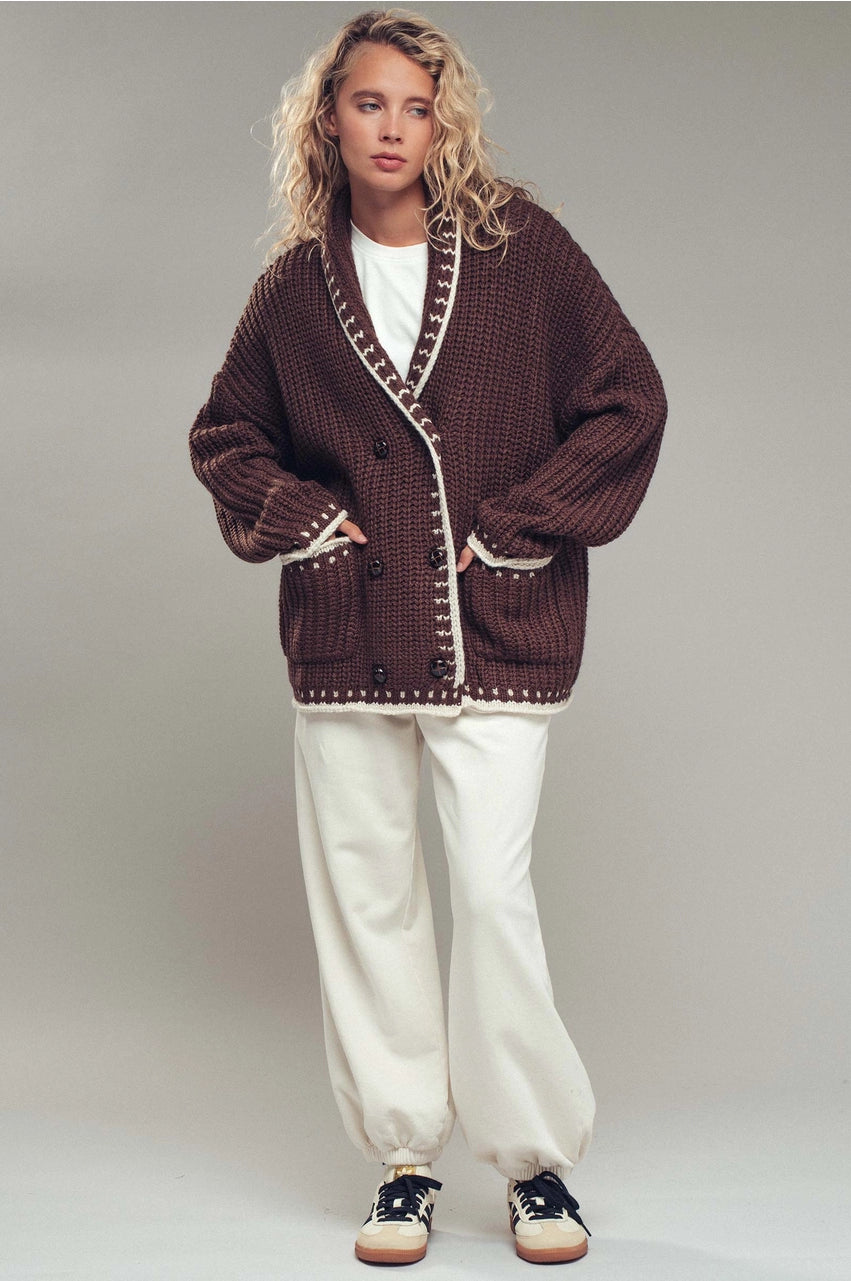 Person wearing a brown knitted cardigan with white trim on a plain background