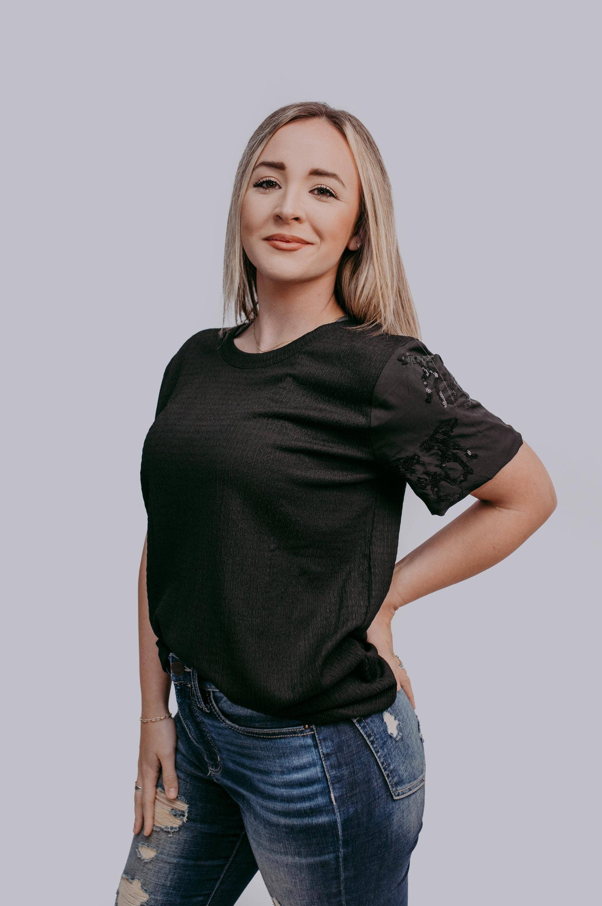 Woman wearing a black top with horse sequin details on sleeves and blue jeans on a plain background
