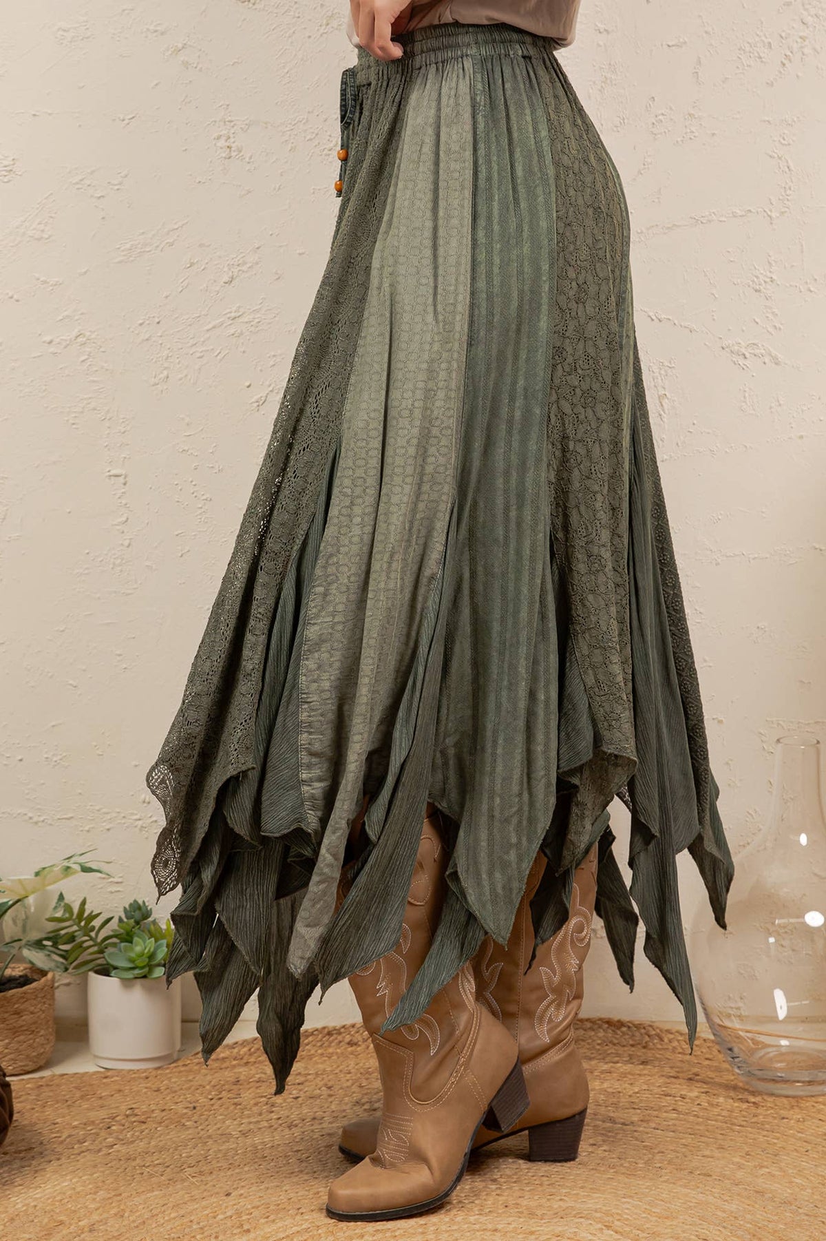 A high-waisted, long pleated skirt in a muted sage green color, styled with brown cowboy boots in a bohemian room setting.