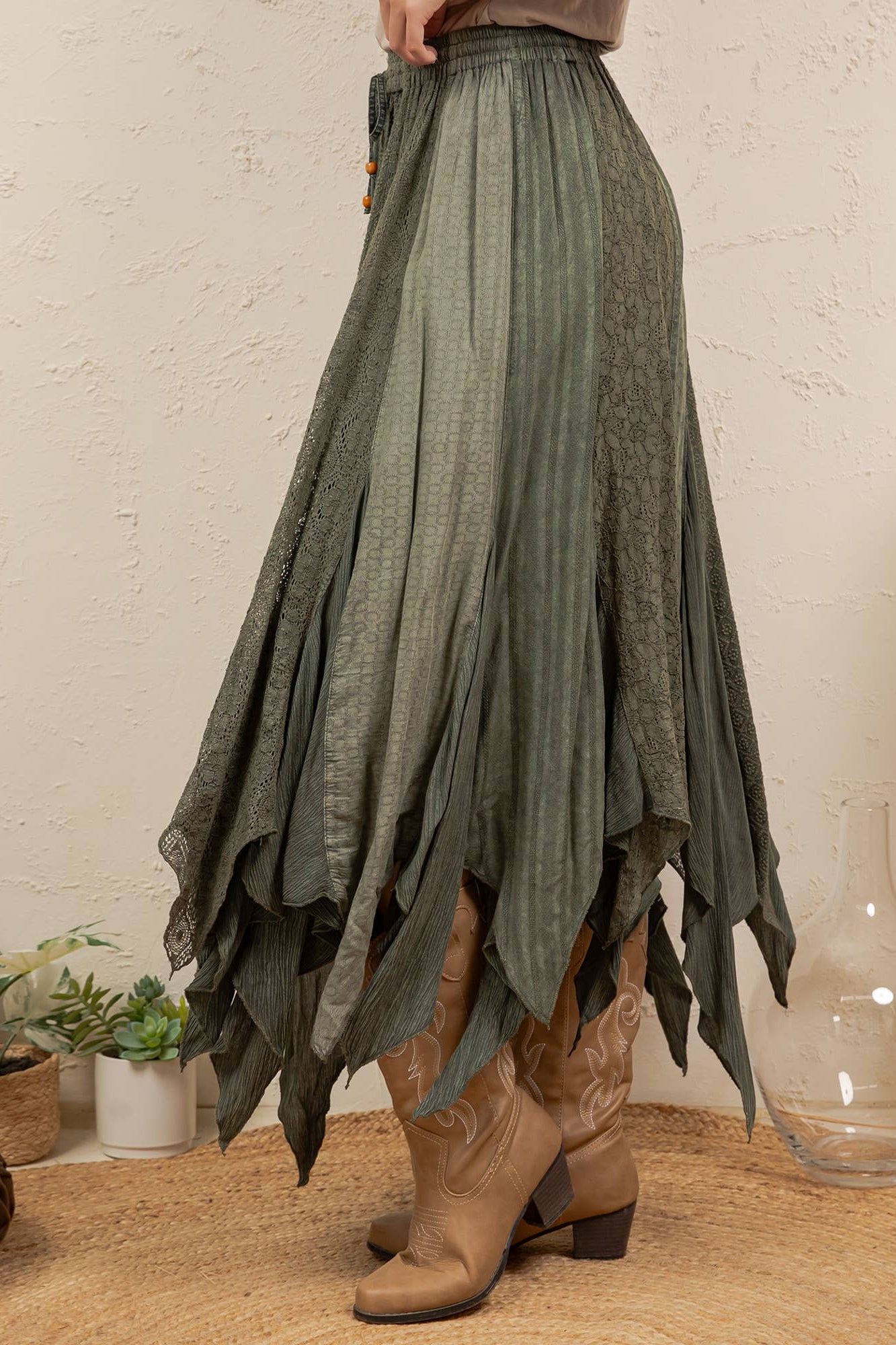 A high-waisted, long pleated skirt in a muted sage green color, styled with brown cowboy boots in a bohemian room setting.