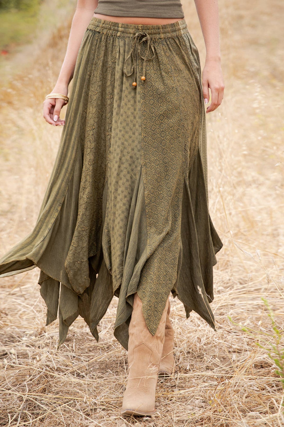 A woman wearing a long, flowing olive green skirt with a tie waist and ruffled asymmetrical hem, walking outdoors in a dry grassy field.