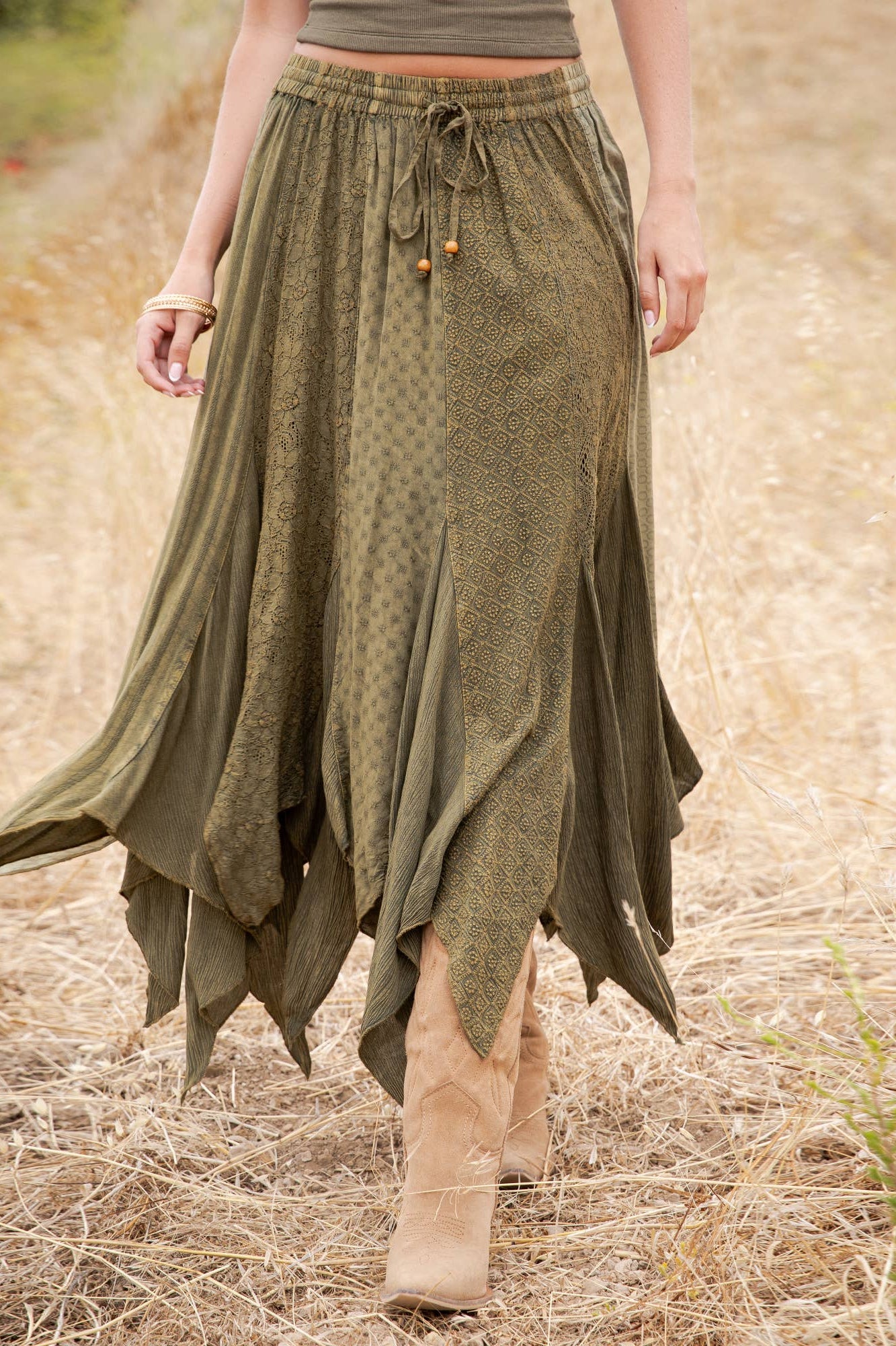 A woman wearing a long, flowing olive green skirt with a tie waist and ruffled asymmetrical hem, walking outdoors in a dry grassy field.