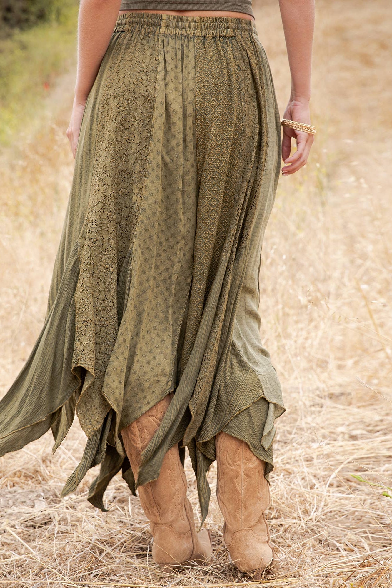 An olive green bohemian maxi skirt with a subtle tonal pattern, shown on a model from behind in a dry grass field. The skirt features an asymmetrical draped hem and is styled with tan cowboy boots.