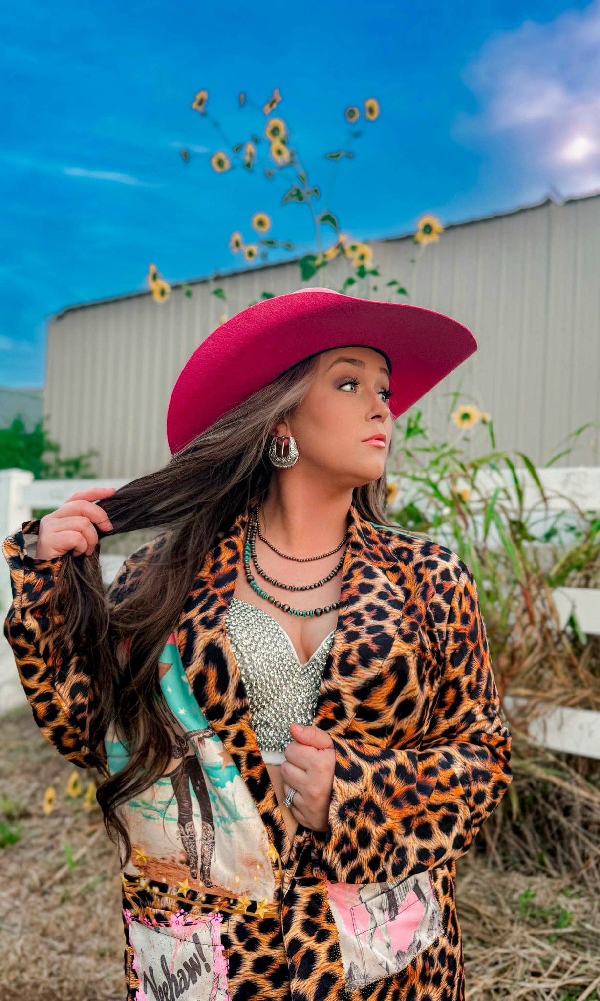 Woman in a leopard print jacket and pink hat standing outdoors with sunflowers and a white building in the background.