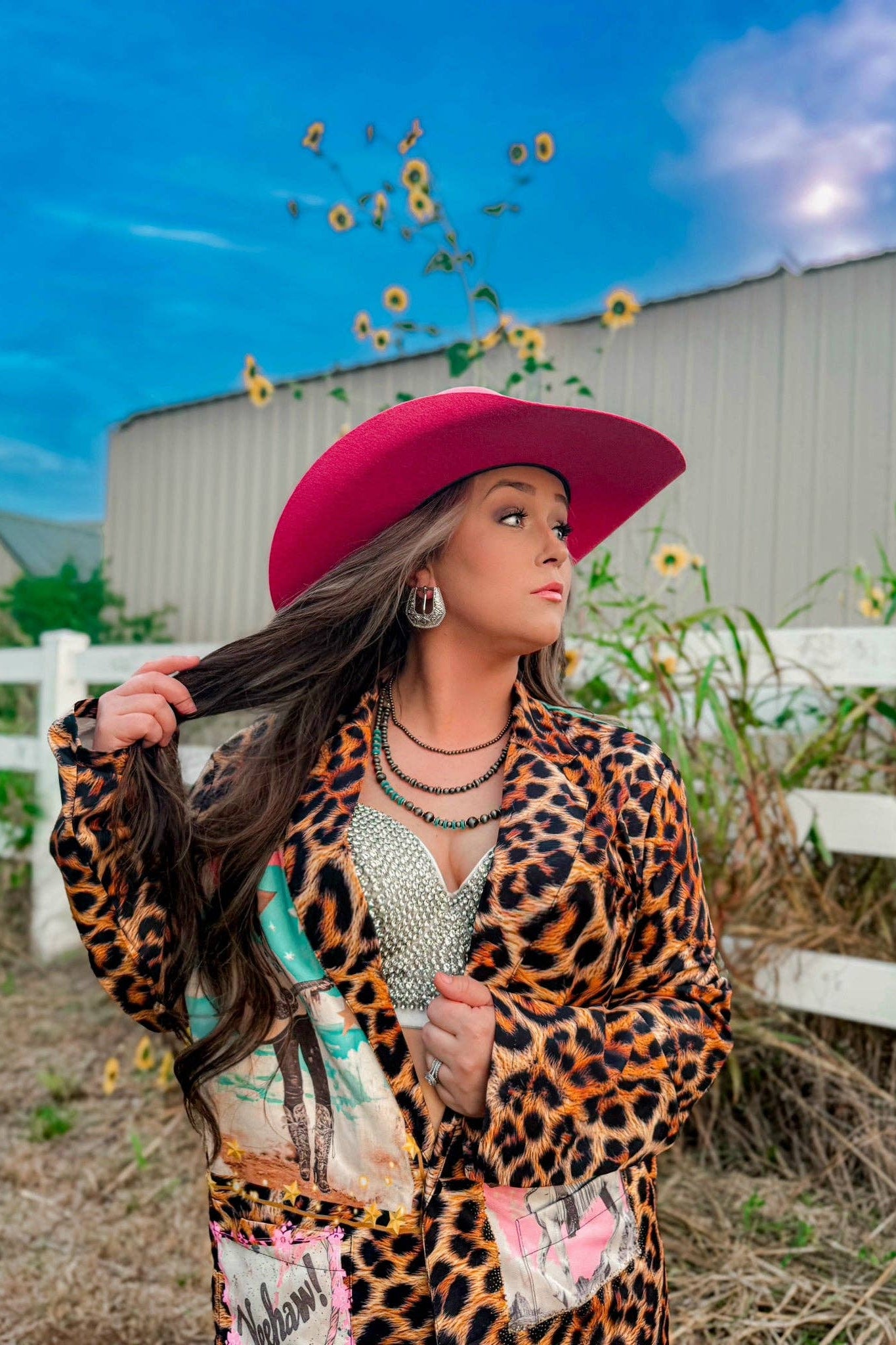 Woman in a leopard print jacket and pink hat standing outdoors with sunflowers and a white building in the background.