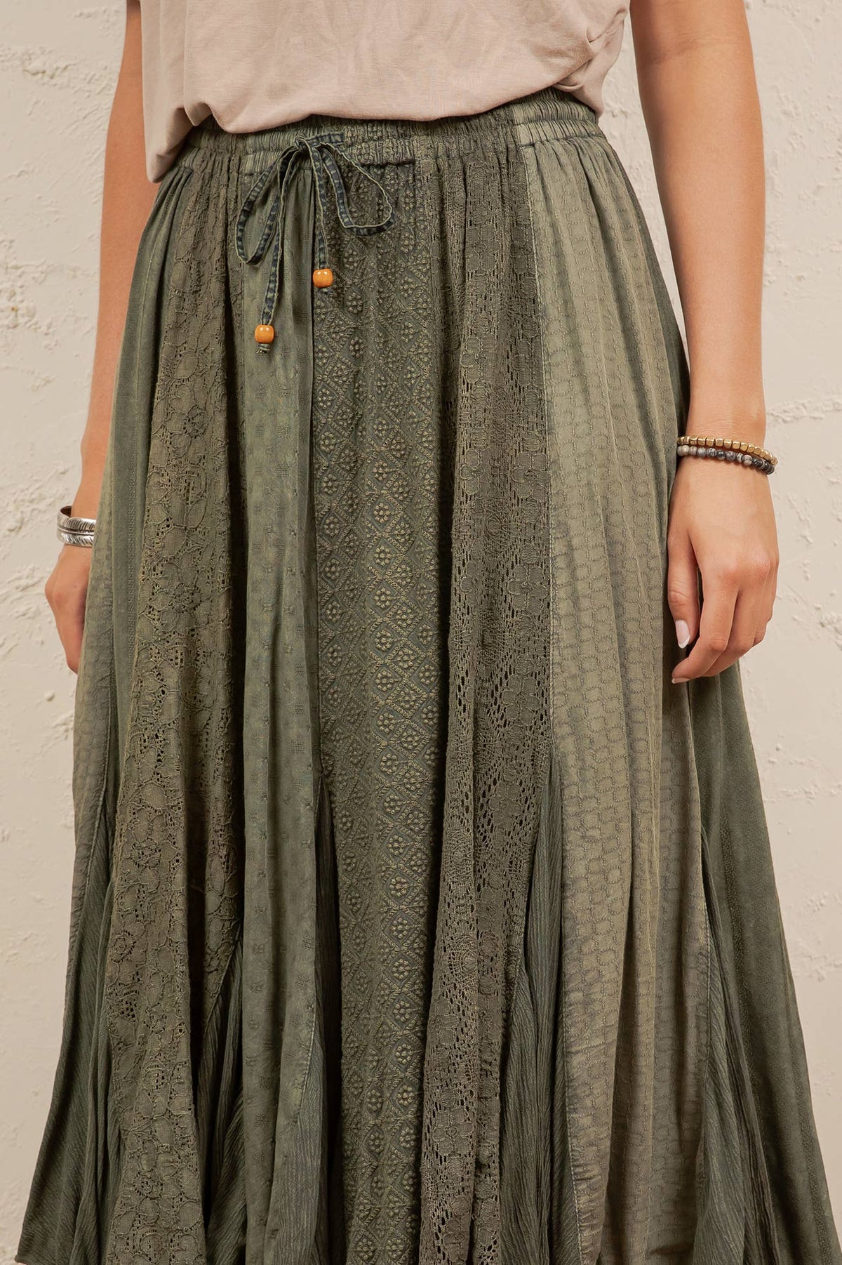 A person wearing a long, pleated skirt in a muted sage green with a subtle tonal geometric pattern. The skirt features an elastic waistband with a drawstring and small orange decorative beads.