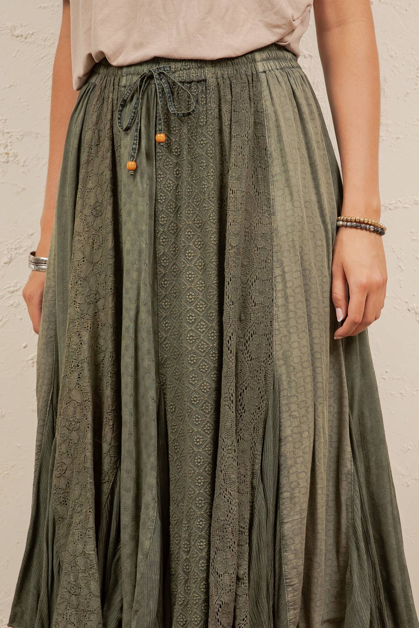 A person wearing a long, pleated skirt in a muted sage green with a subtle tonal geometric pattern. The skirt features an elastic waistband with a drawstring and small orange decorative beads.