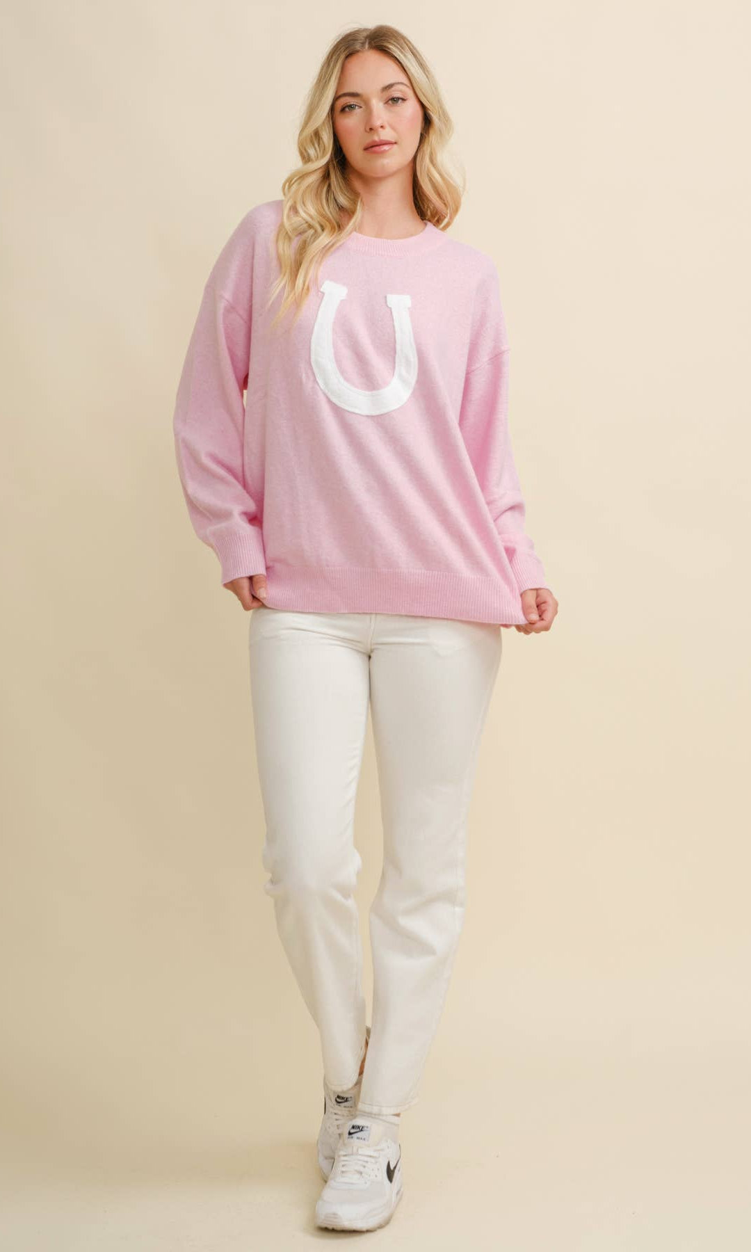 Woman wearing a pink sweatshirt with a white horseshoe and white pants on a beige background