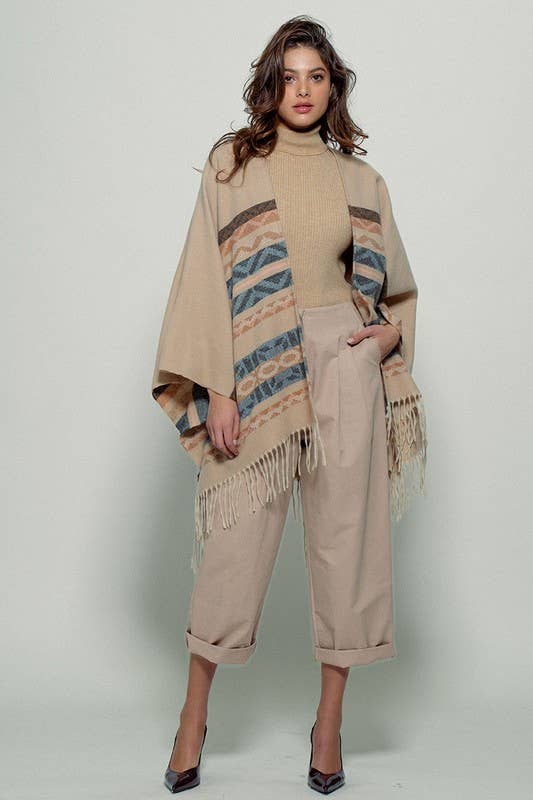 Woman wearing a beige poncho with a Tan background and light blue, brown, light rust, and grey aztec pattern design in stripes with tassel trim along bottom hem. Light weight fabric